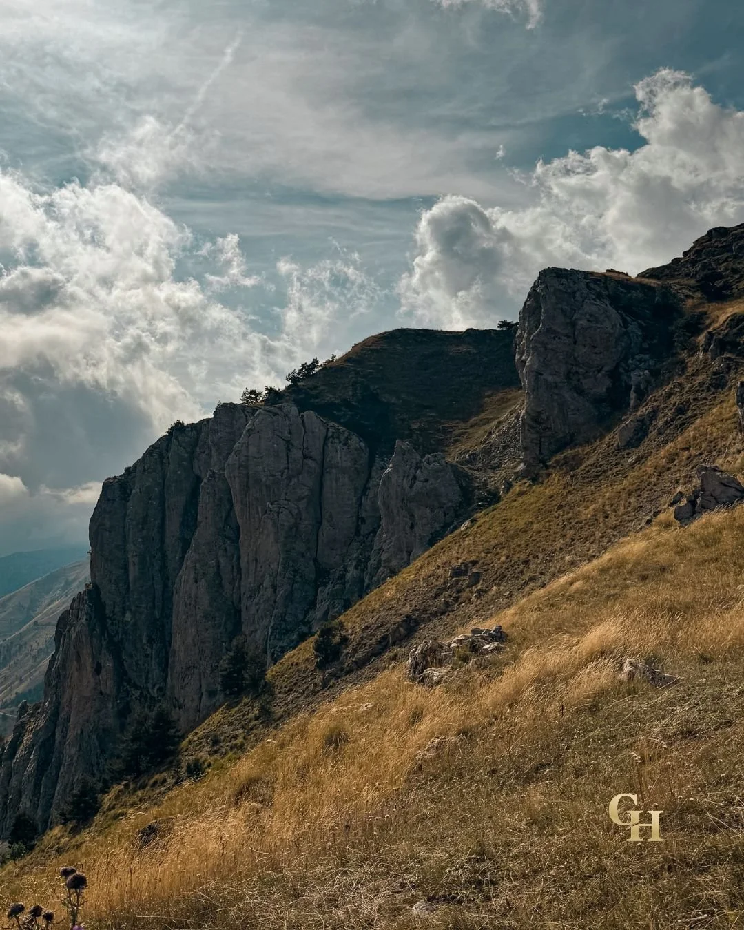 A mountainous landscape with steep rocky cliffs and grassy slopes, under a partly cloudy sky.