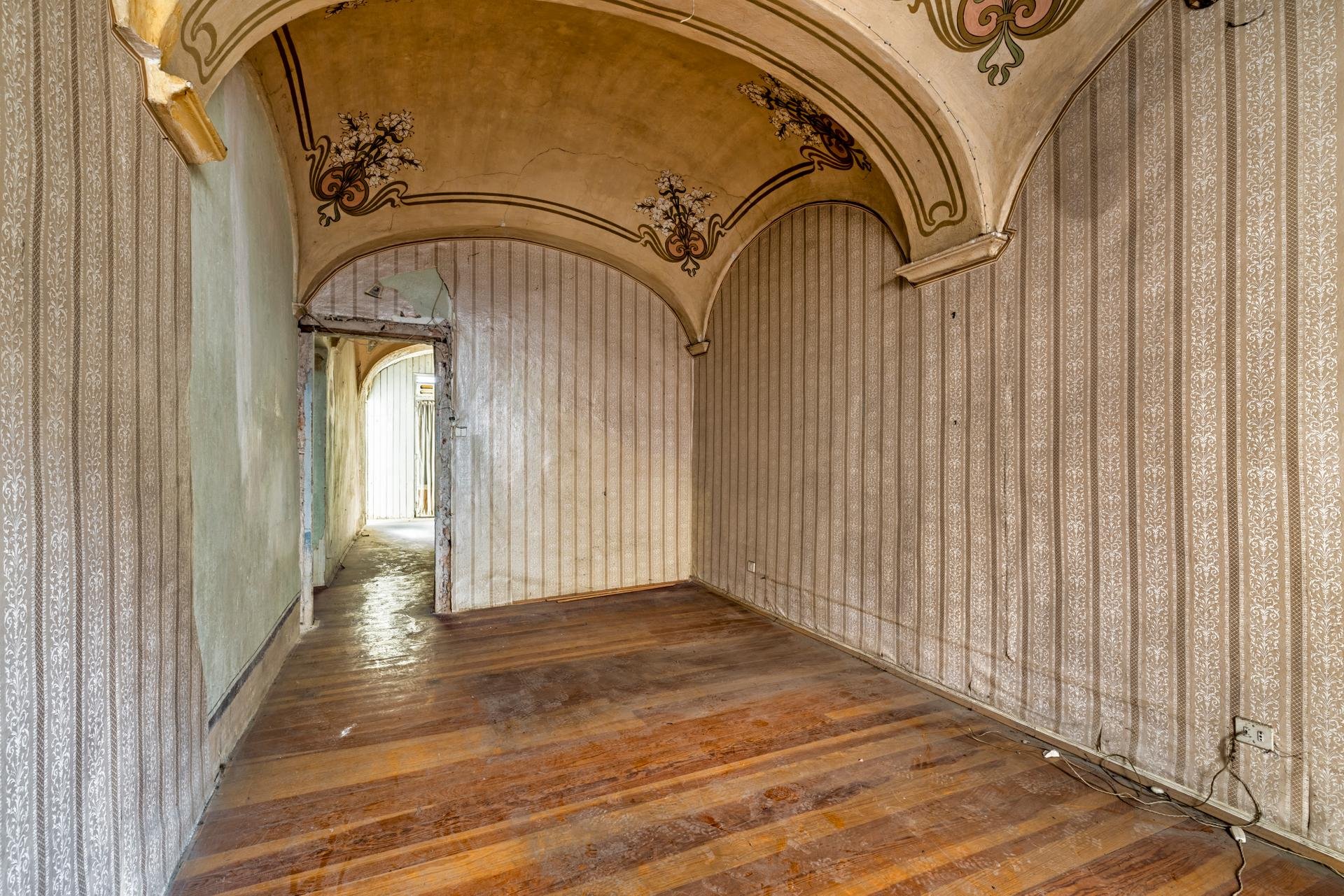 Empty room with worn wooden flooring, decorated wallpaper on the walls, ornate molding and a curved ceiling with floral designs. A doorway at the back leads outside, with visible weathered walls and debris.