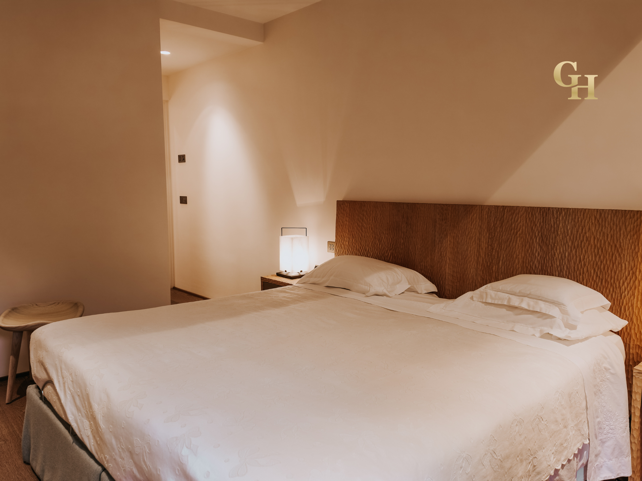 A hotel room with two beds, a wooden headboard, and a bedside table with a lamp. The walls are white, and there is a small lamp on the bedside table. The bed in the foreground has white bedding and pillows.