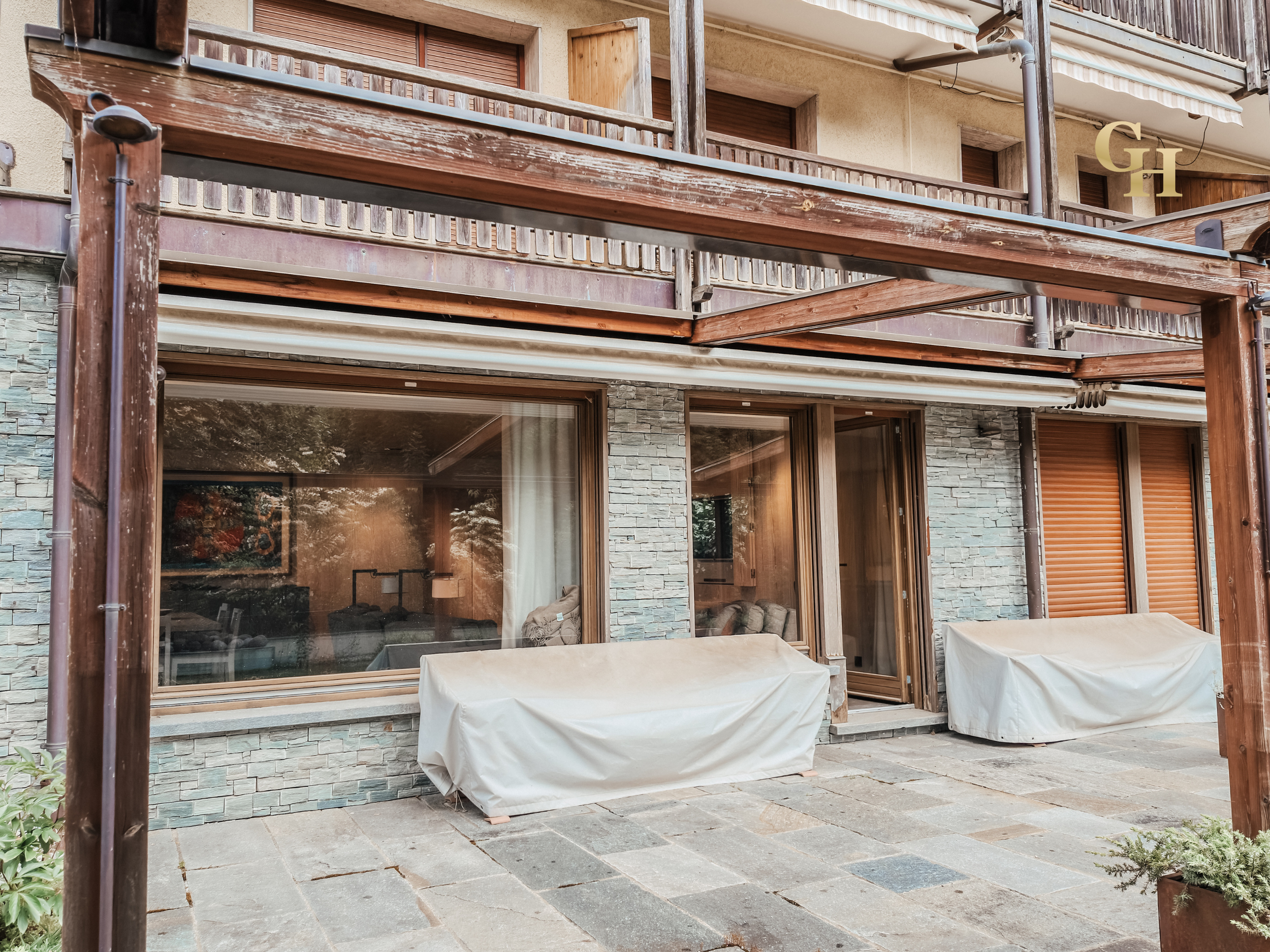 A residential building with large glass windows and a stone facade. A wooden balcony with a weathered finish extends from the upper level. Two outdoor couches, covered with white protective coverings, are positioned on a stone-paved patio below. The 