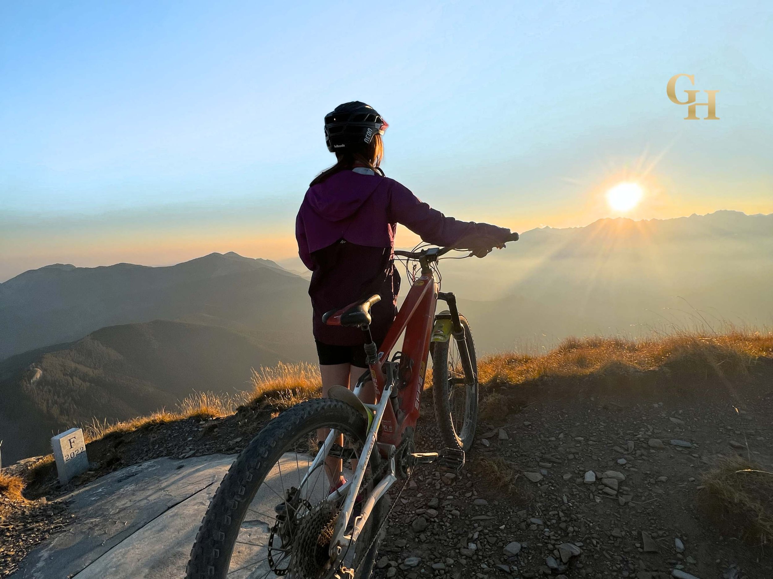 A person with long hair wearing a helmet and a purple jacket standing next to their mountain bike on a trail, overlooking mountains during sunset. The road is Via del Sale, the Salt Road in the Maritime Alps, Italy.
