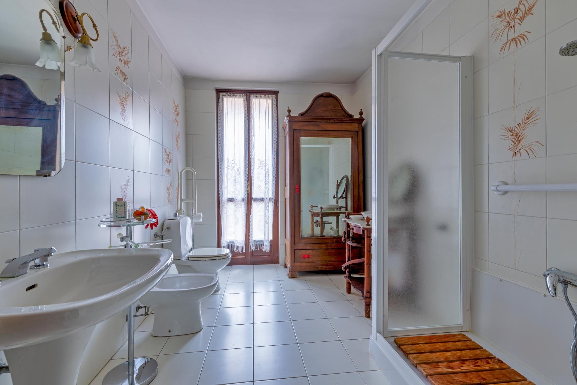 A spacious bathroom with white tiled walls featuring light brown leaf patterns, a window with sheer curtains, a wooden cabinet with a mirror, a shower with frosted glass, a bidet and sink, and various bathroom accessories.