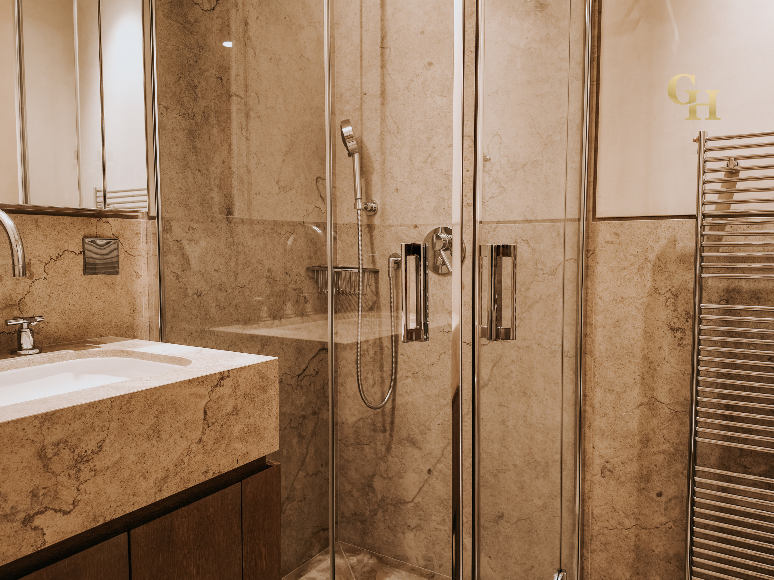 Interior of a modern bathroom with a glass shower enclosure, beige marble walls, a towel warmer, and a vanity with a stone countertop.