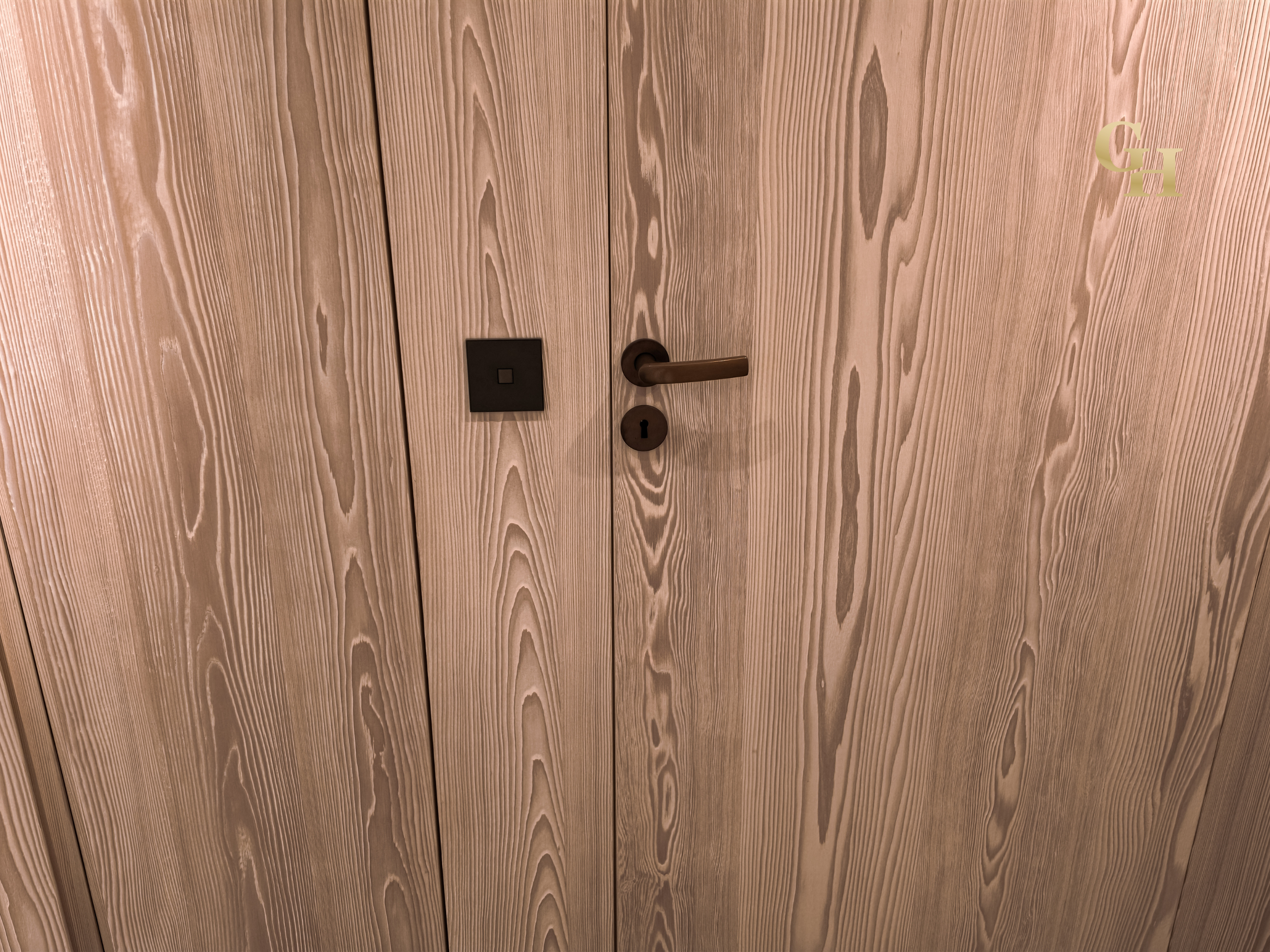 A wooden door with a modern door handle and a lock, featuring a small square doorbell or button near the handle. The door has a natural wood grain pattern, with some letters 'CH' visible in the upper right corner.