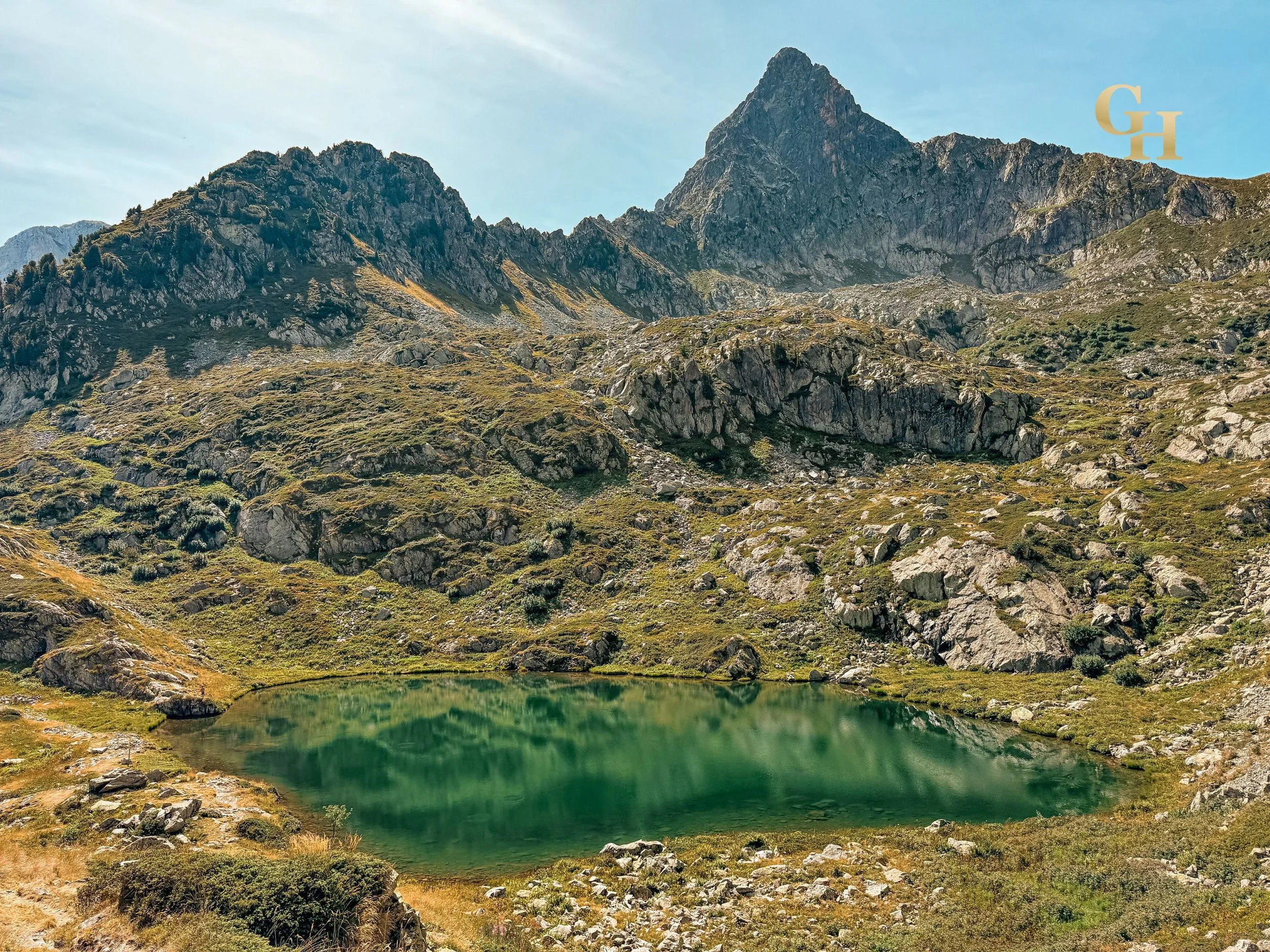 A mountain with a peak and rocky terrain, a small green lake at its base, surrounded by grassy and rocky landscape under a partly cloudy sky.