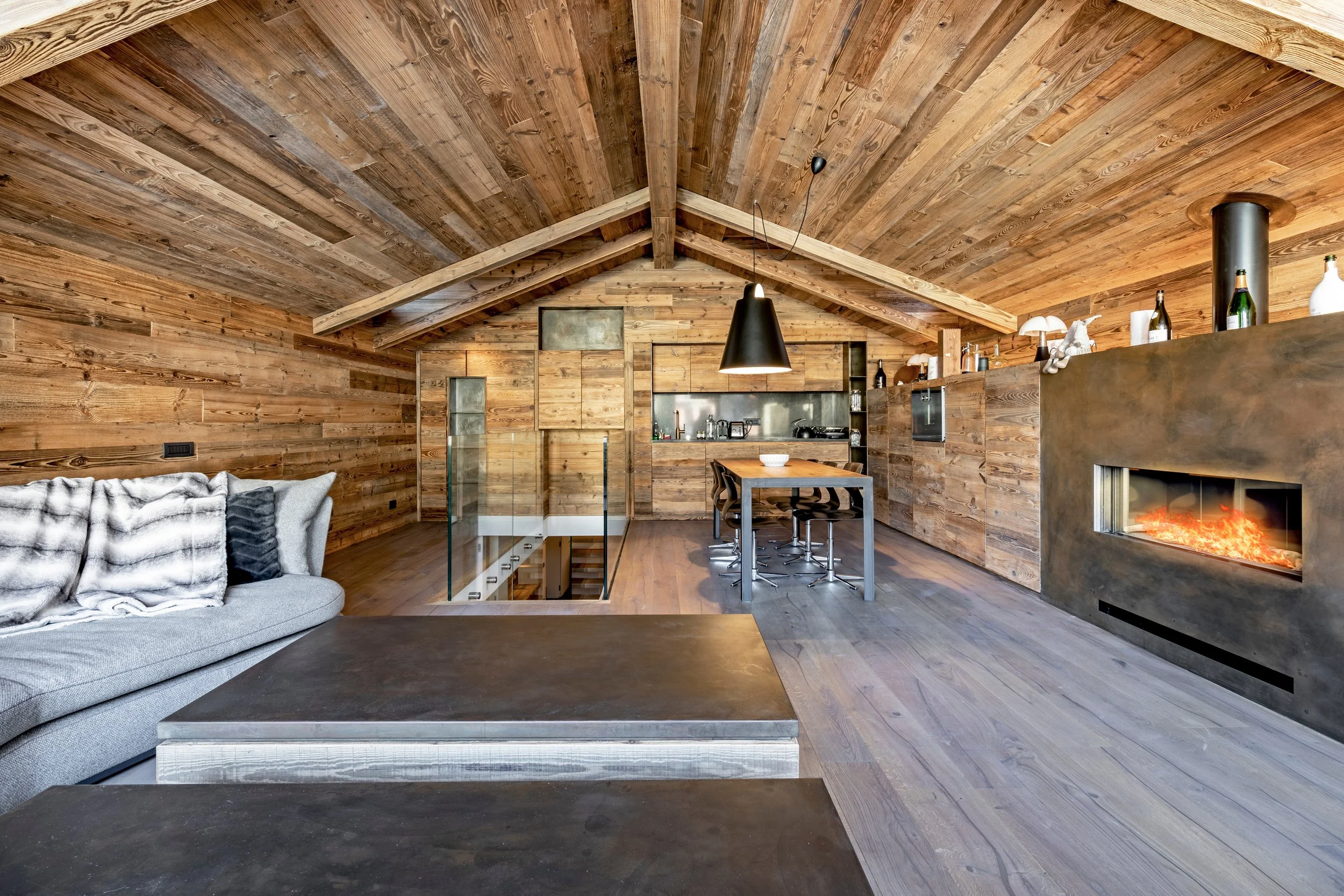 A living room with wooden walls and ceiling, a gray sofa with throw pillows, a black coffee table, a dining table with black chairs, a black hanging light fixture, a fireplace, and kitchen area in the background.