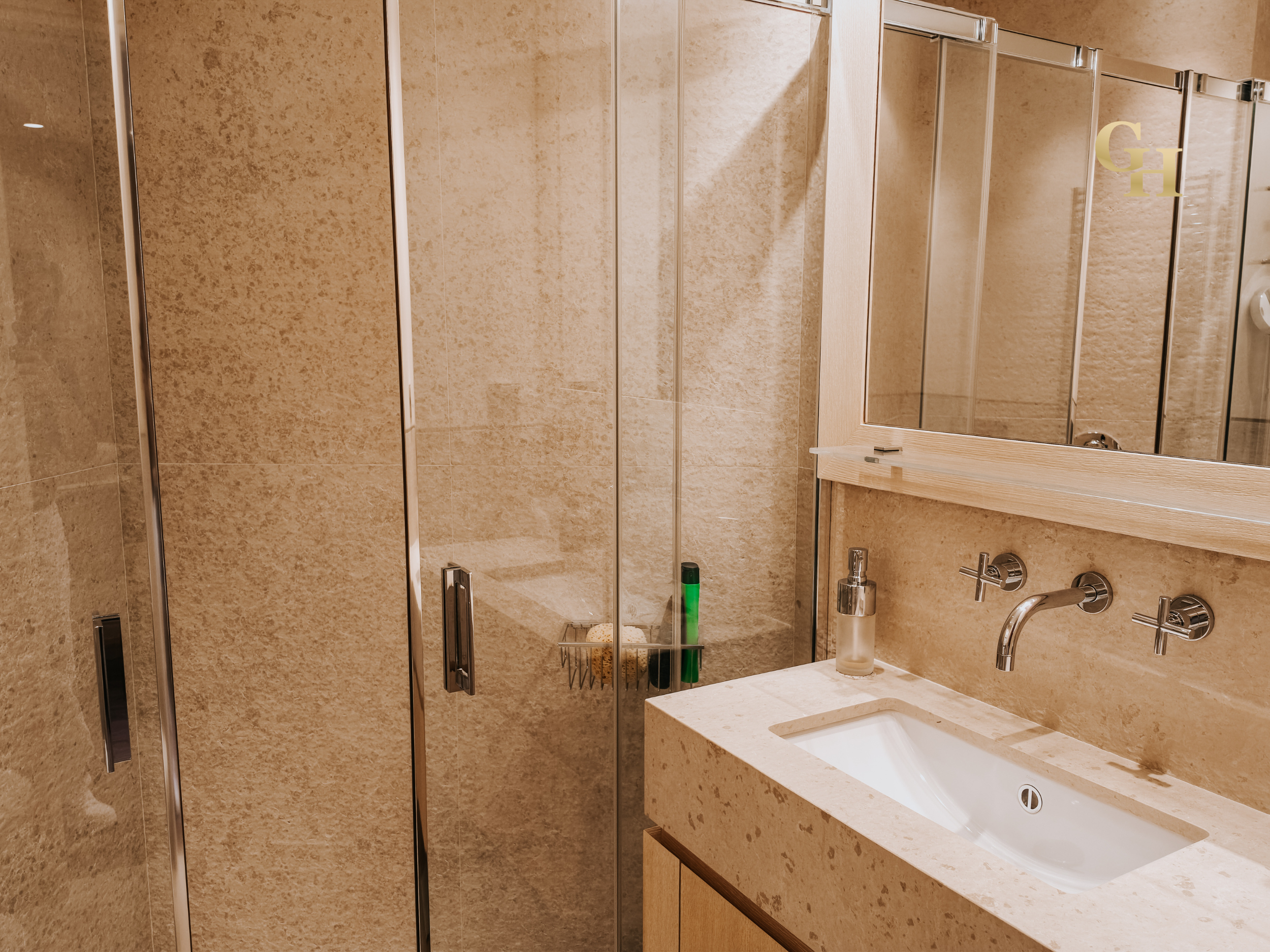 Bathroom with beige tiled walls, a mirror, a sink with a faucet, a soap dispenser, and a shower with glass doors.