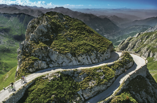 A narrow winding mountain road carved into the rocky terrain, with bicycles riding along it, surrounded by green hills and distant mountain peaks under a partly cloudy sky.