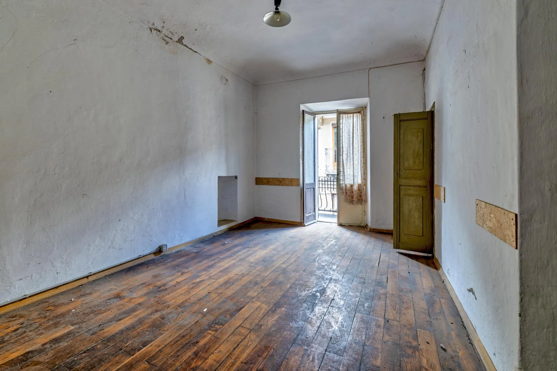 Empty room with worn hardwood floor, white textured walls with cracks, a doorway with partially open double doors leading outside, and a single ceiling light fixture.