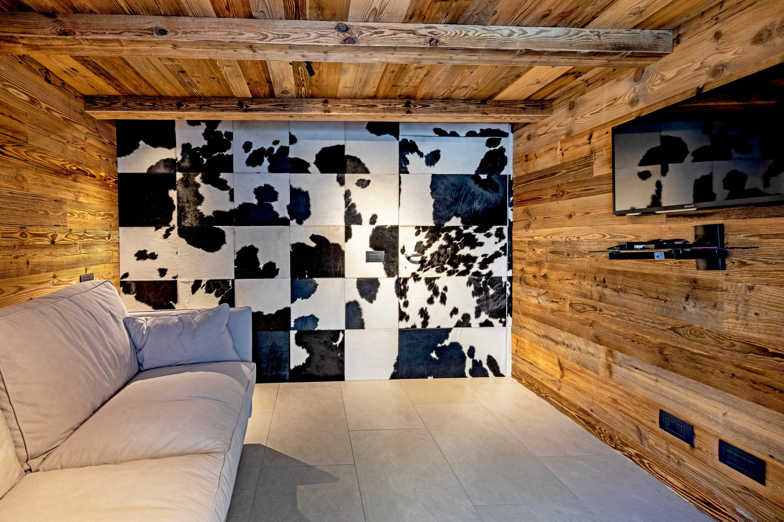 A living room corner with a beige couch, wooden wall and ceiling, a cow-print rug, and a wall-mounted television.