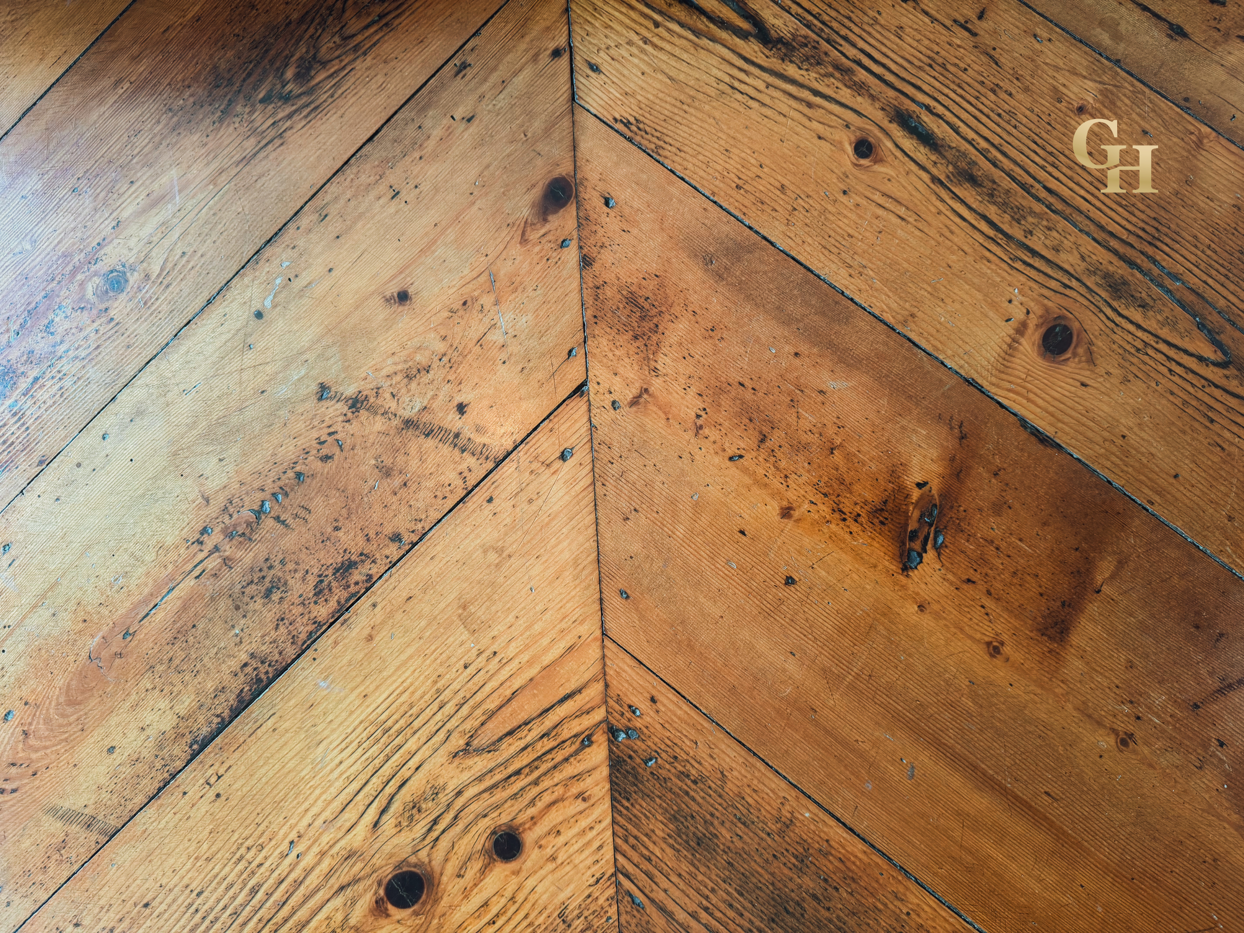 Close-up of a wooden floor with a chevron pattern, showing natural wood grain, knots, and some small paint spots.