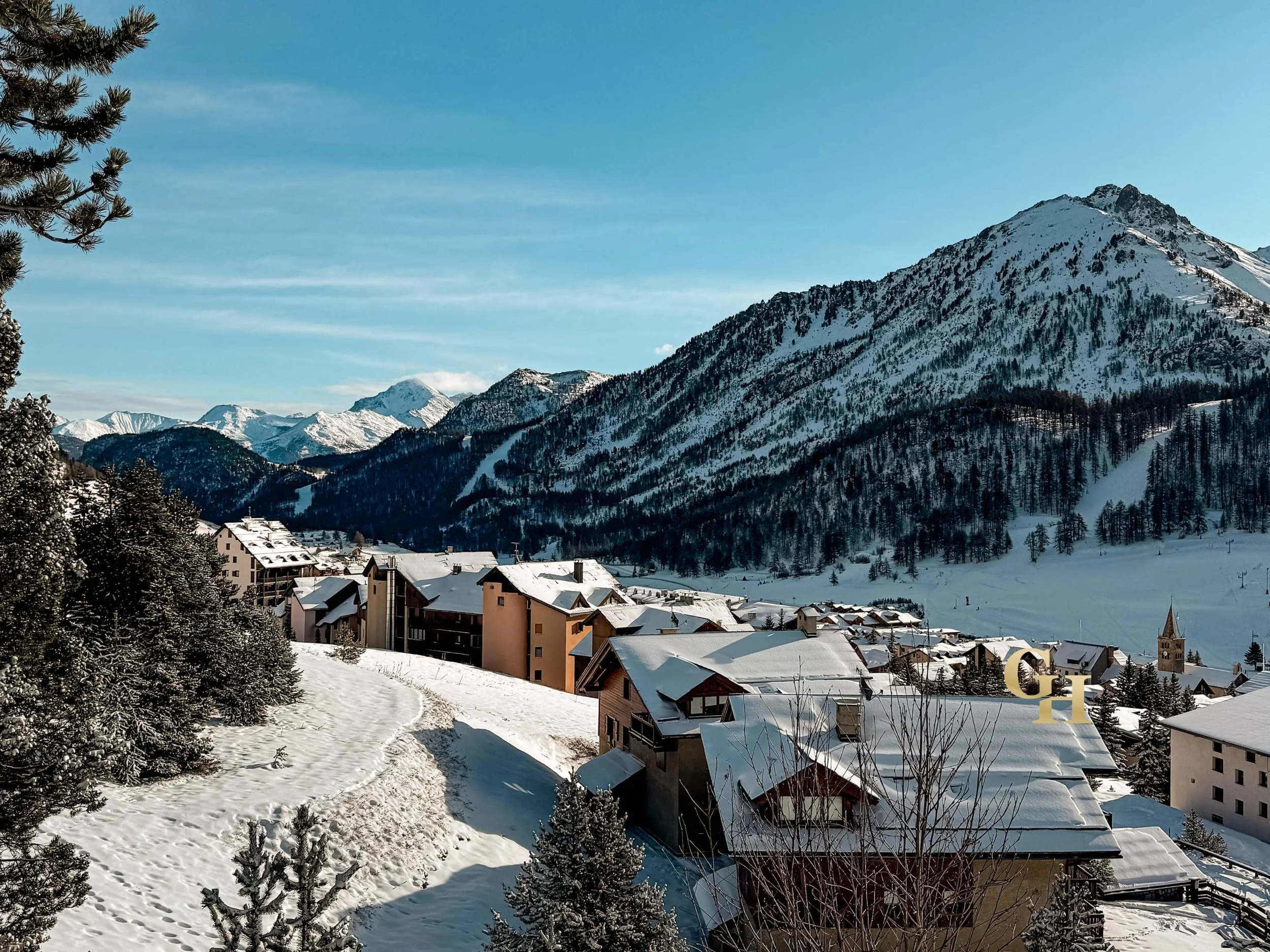Snow-covered mountain village with residential buildings and a church, surrounded by snow and trees, under a clear blue sky.