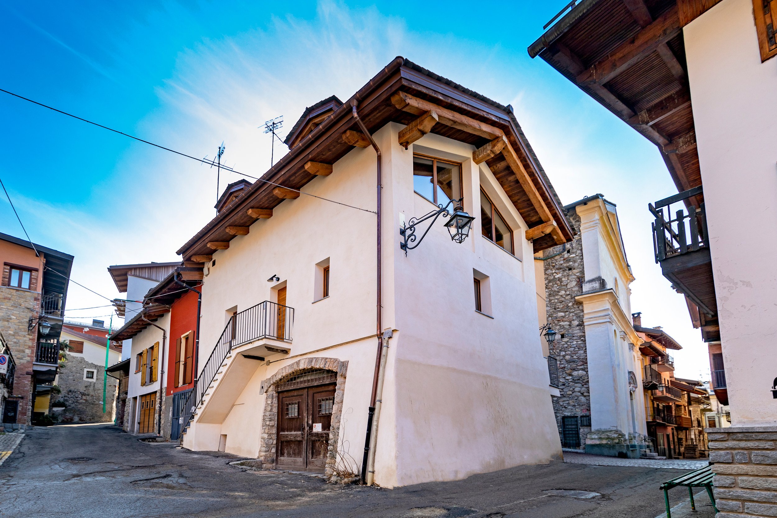 View of European-style buildings with stucco and stone facades, wooden window frames, and balconies on a narrow street under a clear blue sky.