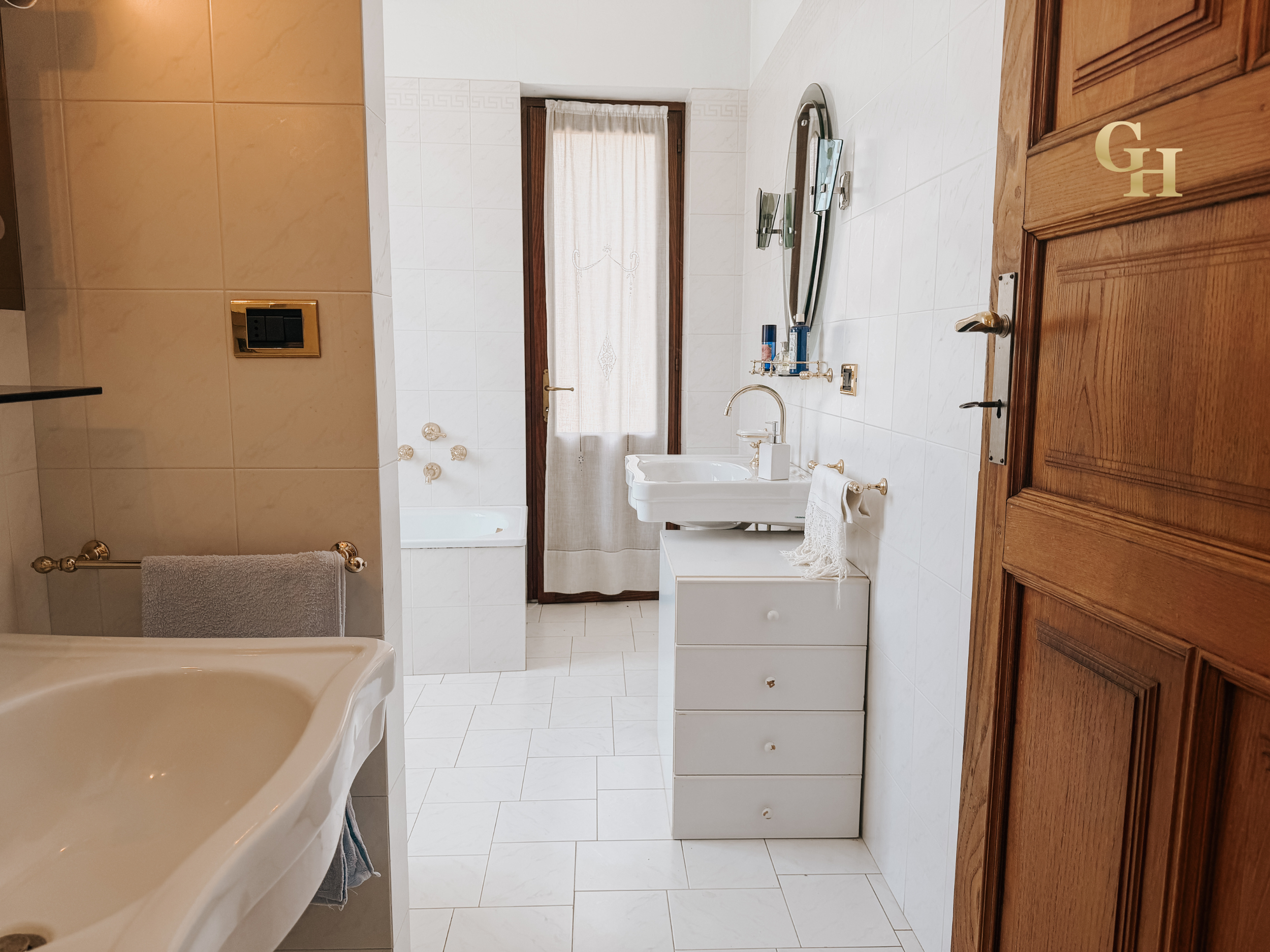 A bathroom with white tiled walls and floor, wooden door, and a window with a curtain. There is a sink with a mirror above it, some shelves, and a towel hanging on a gold towel rack.