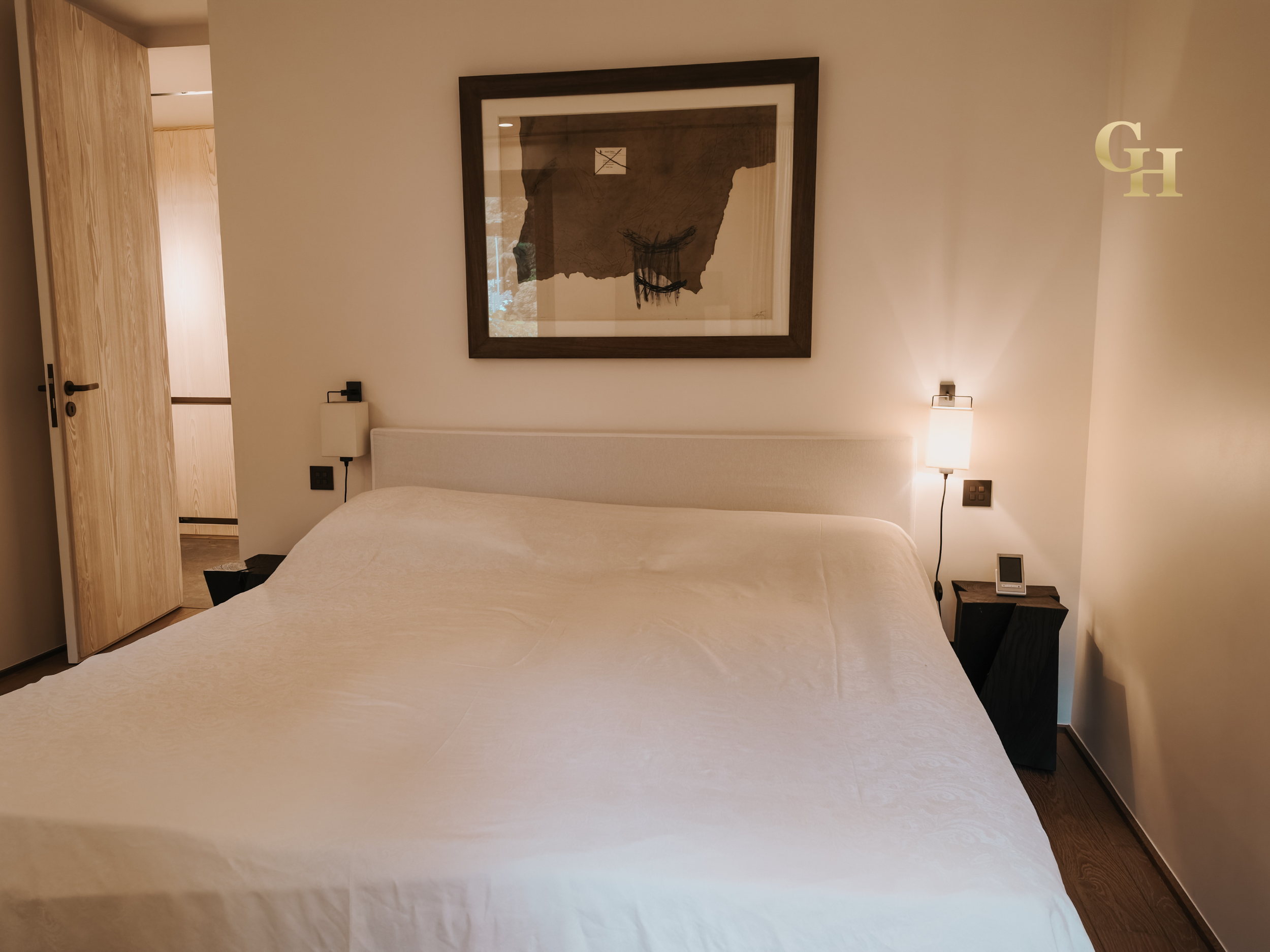 A hotel room with a bed, framed artwork on the wall, two black nightstands with lamps, a digital clock, and visible doorways.