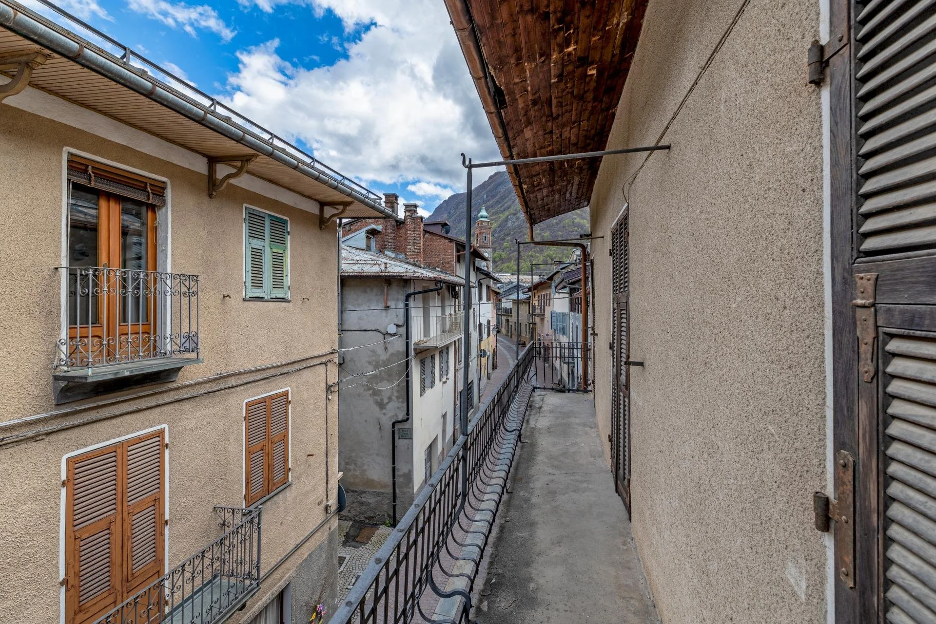 Narrow balcony overlooking a street in a mountainous town, with buildings featuring wooden shutters and balconies, under a partly cloudy sky.