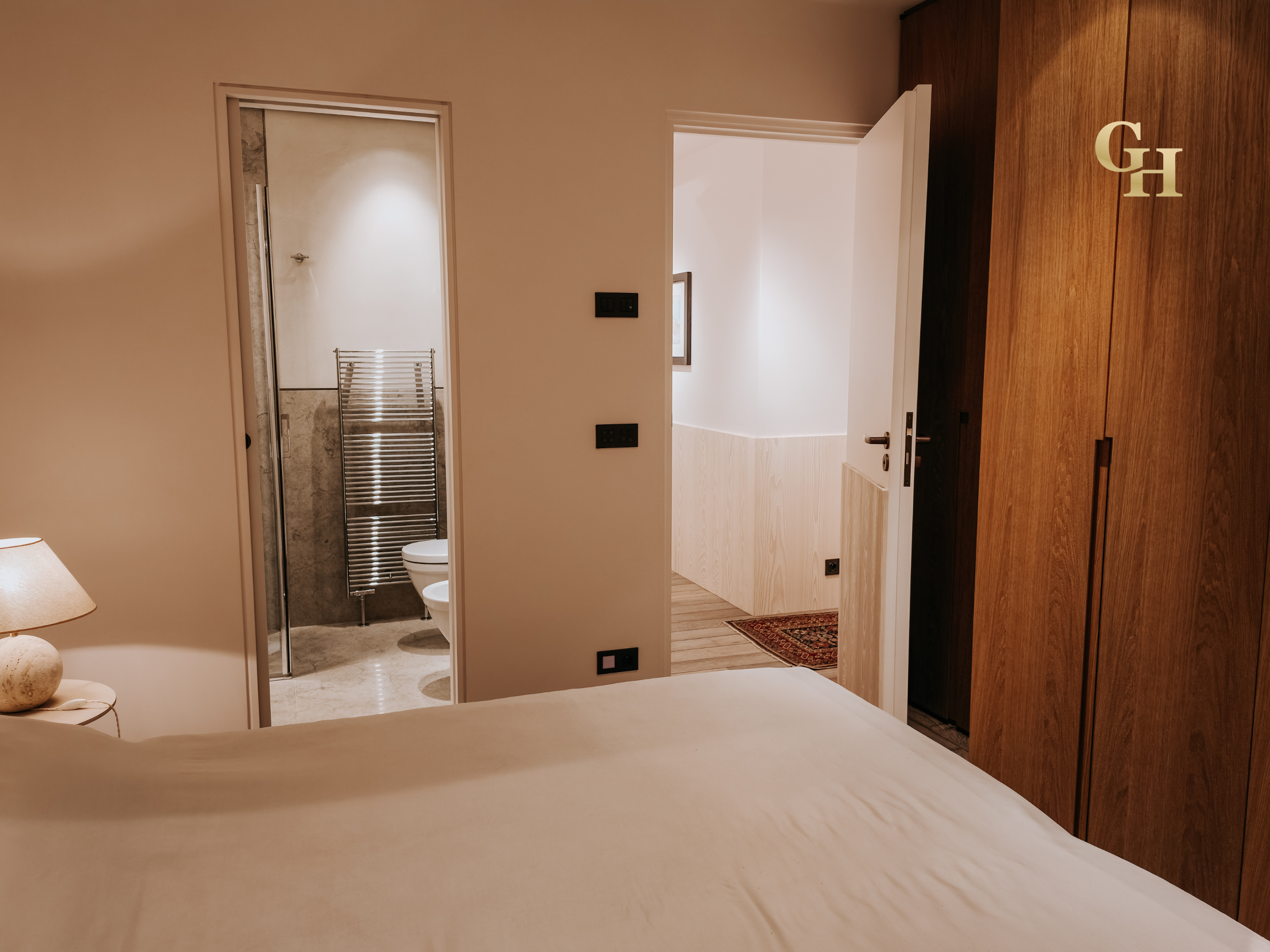 A view from a bedroom looking into a bathroom. The bathroom has a glass shower enclosure, a heated towel rail, and a toilet. The bedroom has a wooden wardrobe, a lamp, and a bed with white bedding.