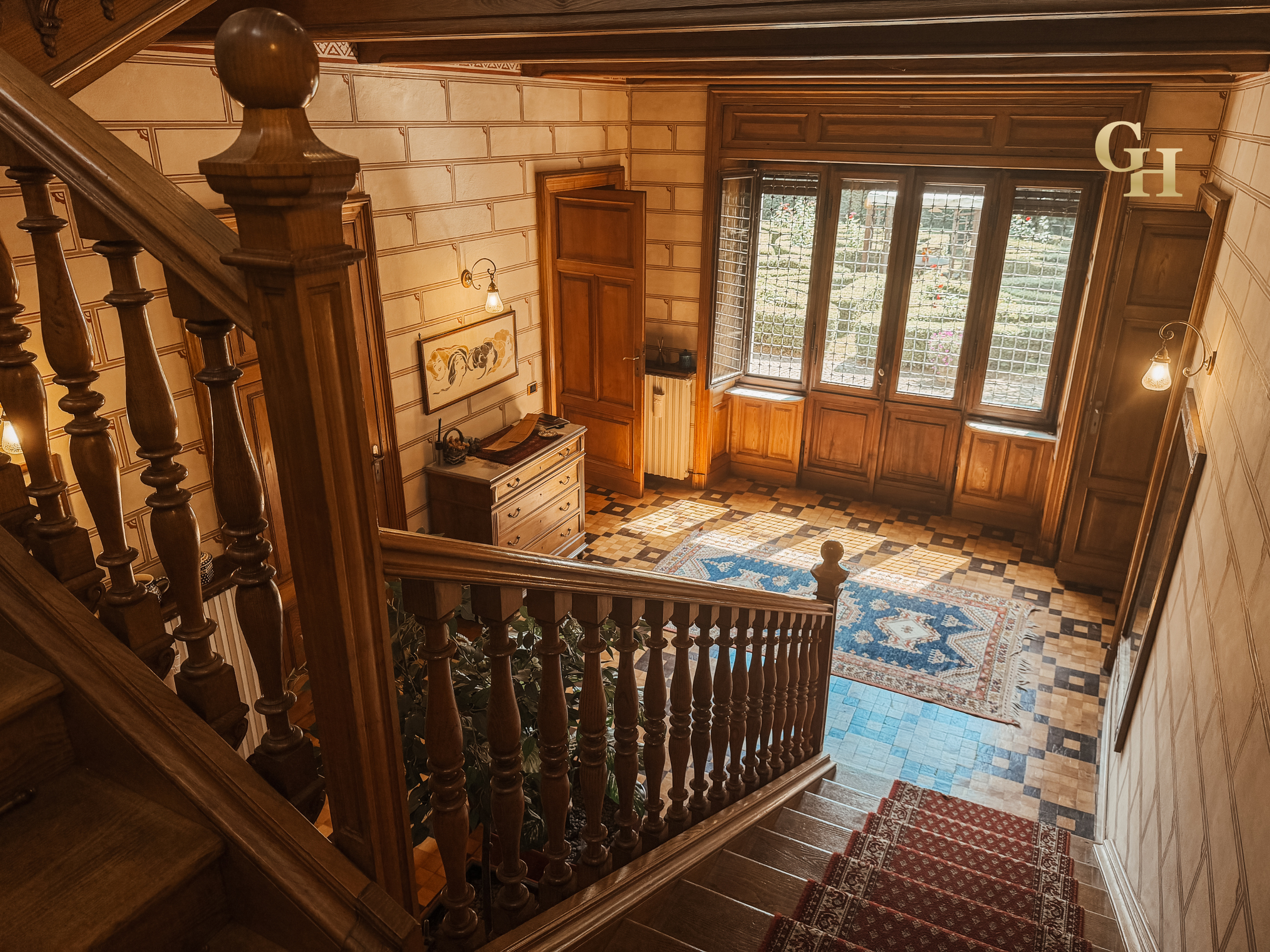 View from the top of a wooden staircase looking down into a foyer with large windows, wooden walls, and a patterned carpet on the stairs and floor.