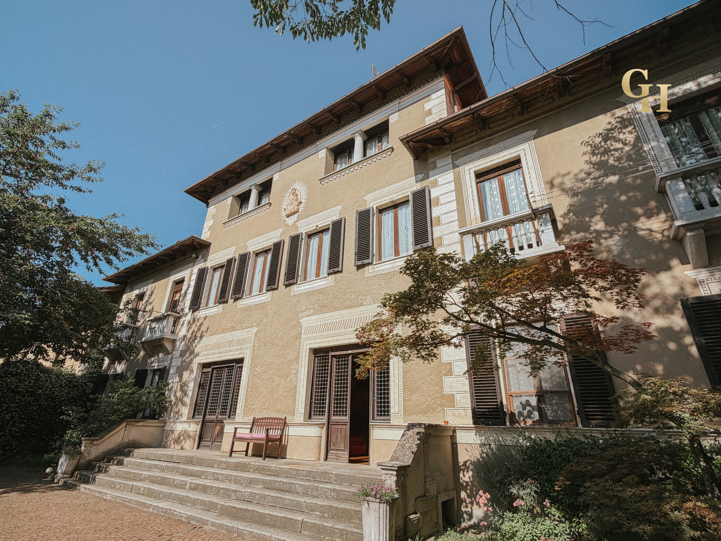 A multi-story building with beige exterior walls and dark window shutters, surrounded by trees with green leaves, under a clear blue sky.