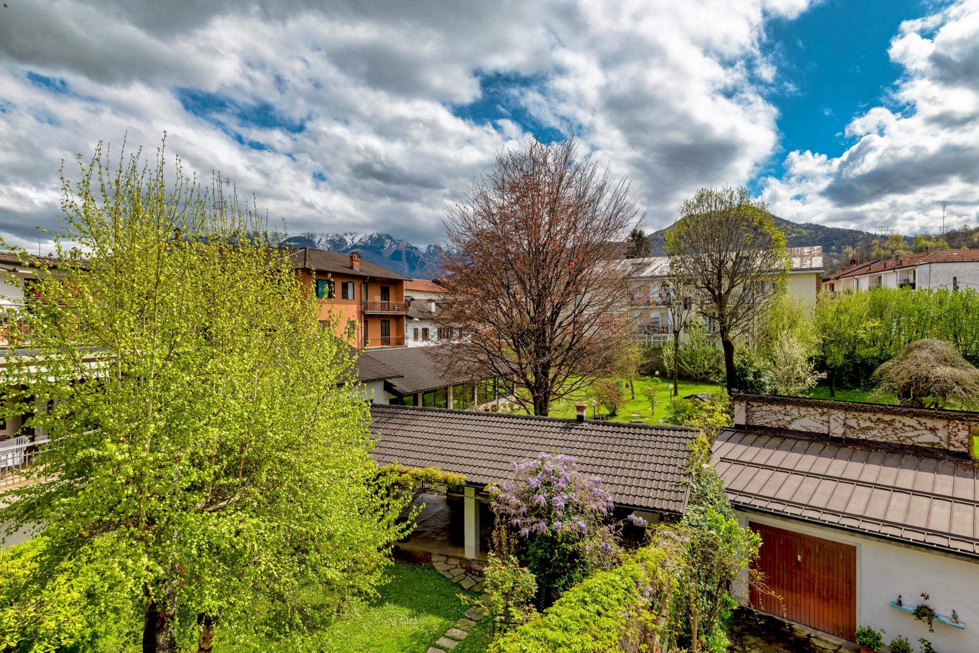 A scenic view of a neighborhood with houses, trees with spring foliage, and mountains with snow in the background under a partly cloudy sky.