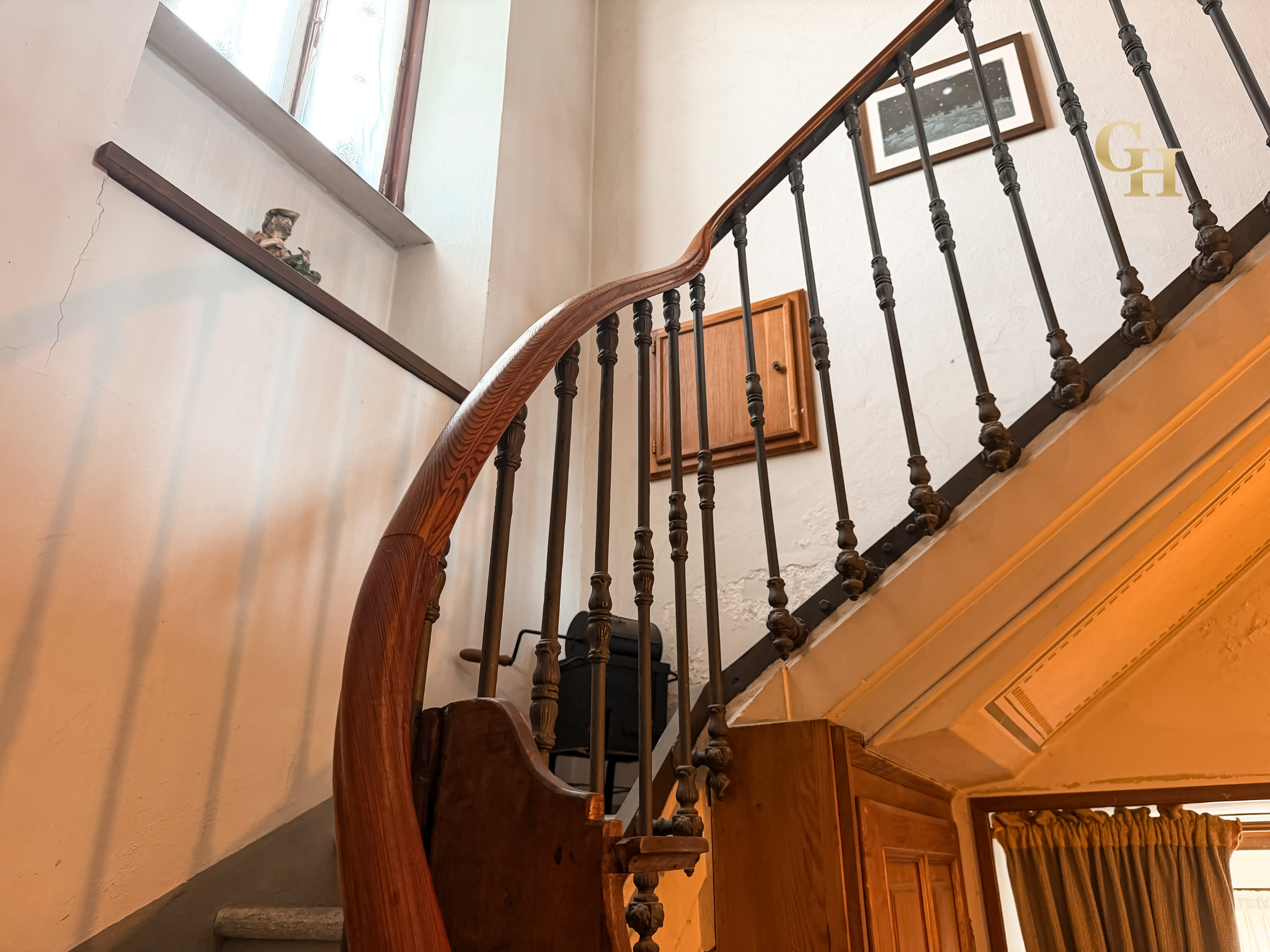 Interior view of a staircase with a wooden handrail and black metal balusters, a framed picture on the wall, and a small window allowing natural light into the space.