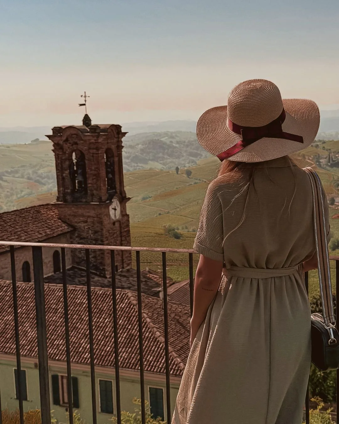 A woman in a beige dress and large straw hat stands on a balcony overlooking an Italian hillside town with a church tower and rolling vineyards in the background.