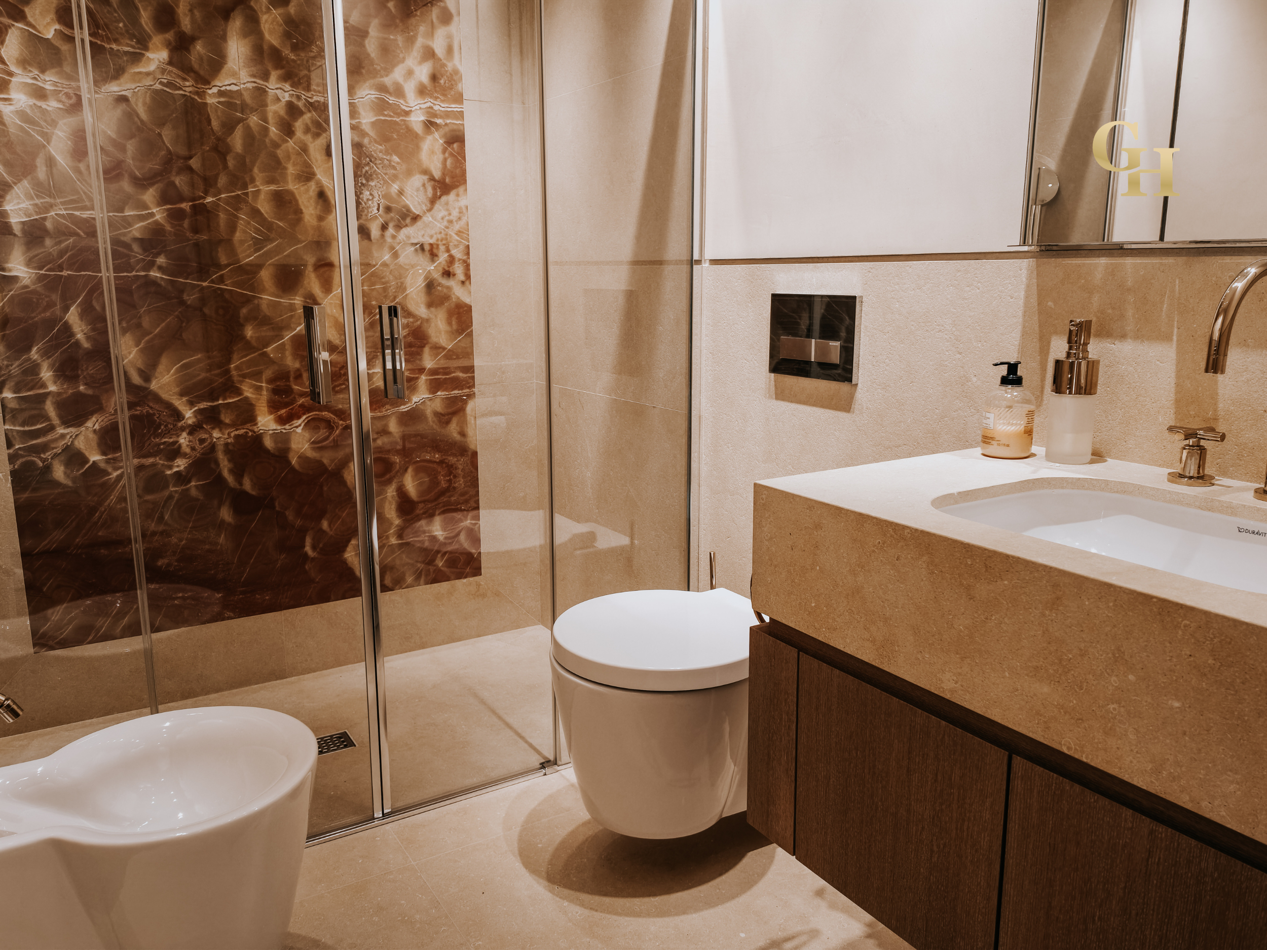 A modern bathroom with marble and beige tile walls, a glass shower enclosure with metal handles, a white toilet, a beige sink with a wooden cabinet, and various soap dispensers on the countertop. There's a large mirror above the sink with the initial