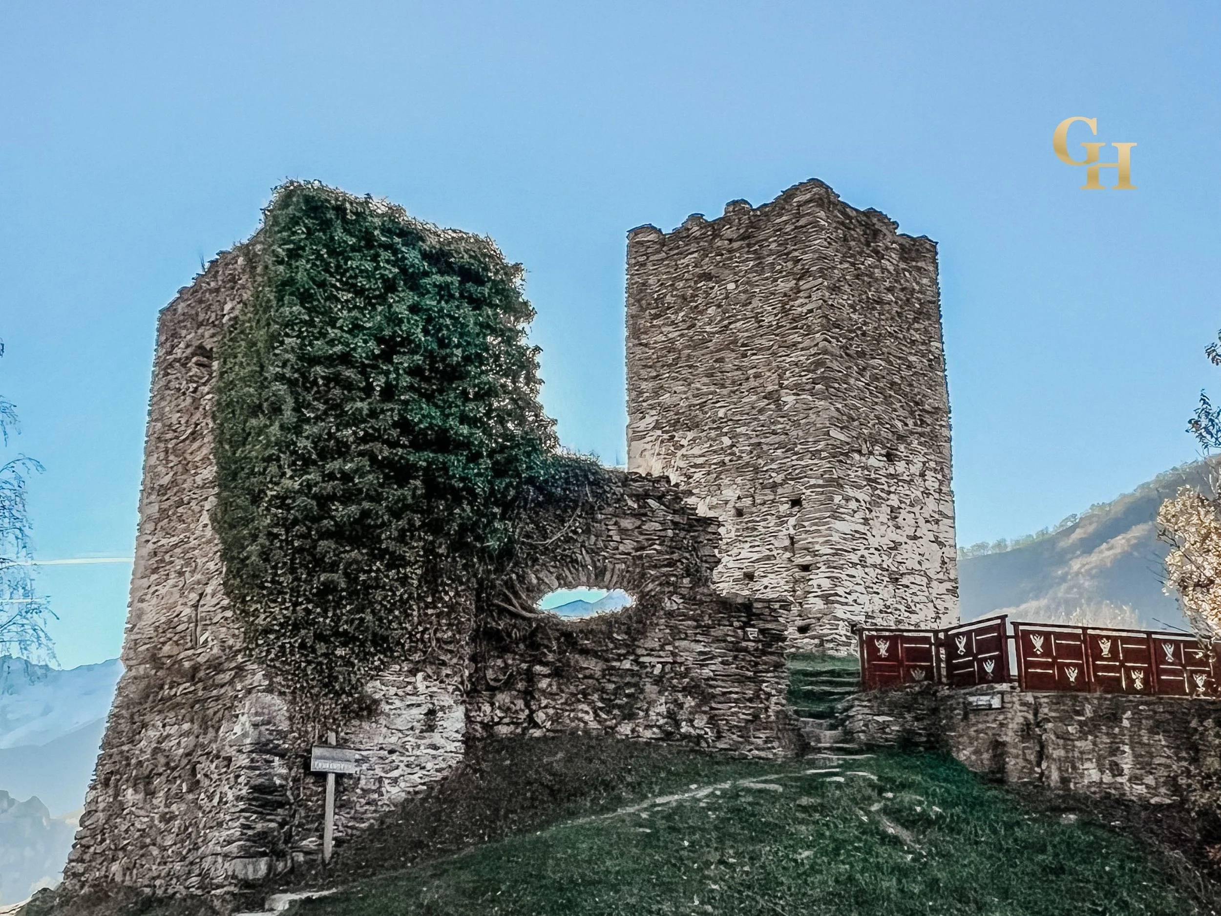 Ruins of an ancient stone structure with a large central arch and two tall towers made of stacked stones, surrounded by trees and greenery, under a clear blue sky.