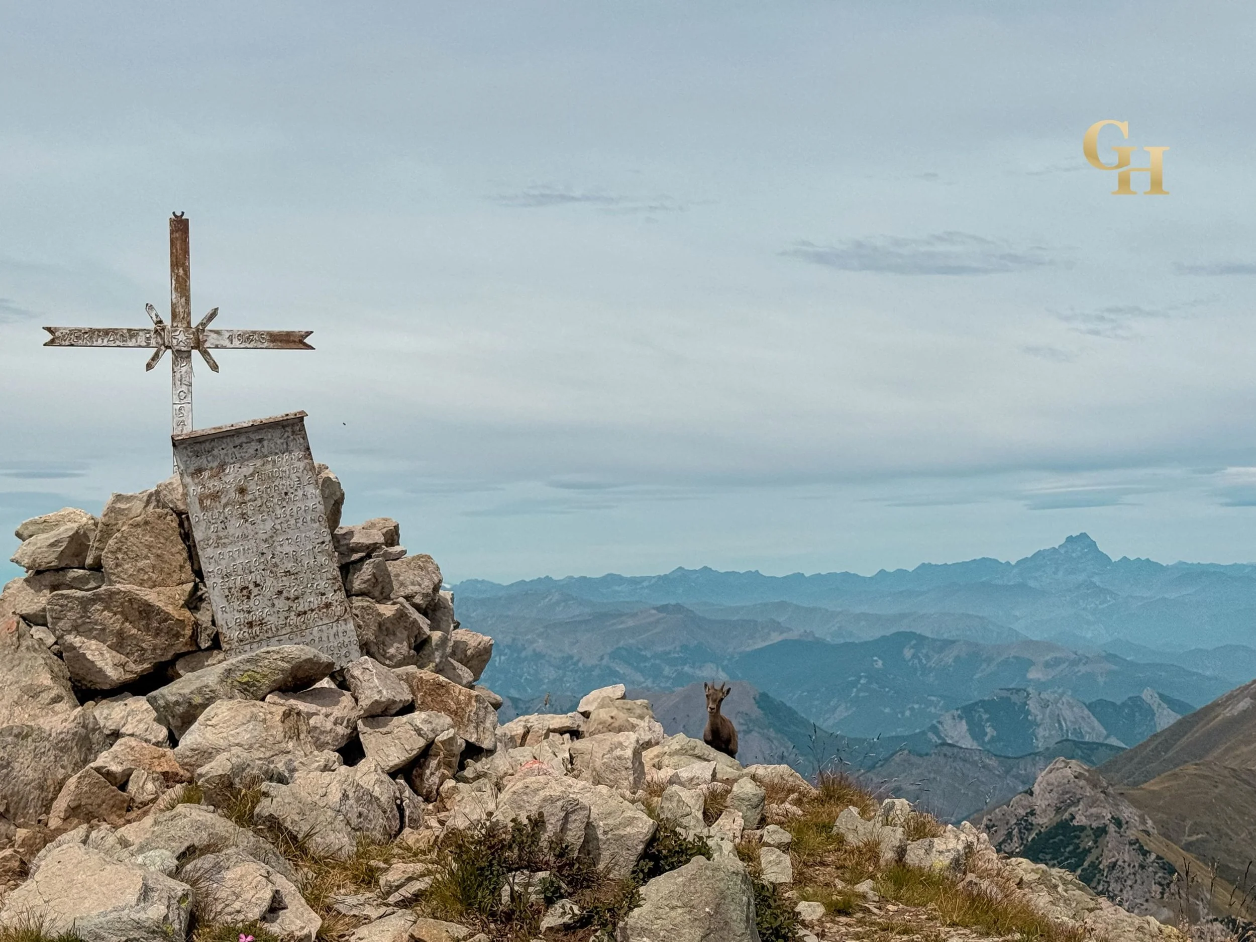 Mountain trail with rocks, a small placard and a cross perched on rocks, with a mountain goat standing on rocky terrain and mountains in the distance under a partly cloudy sky.
