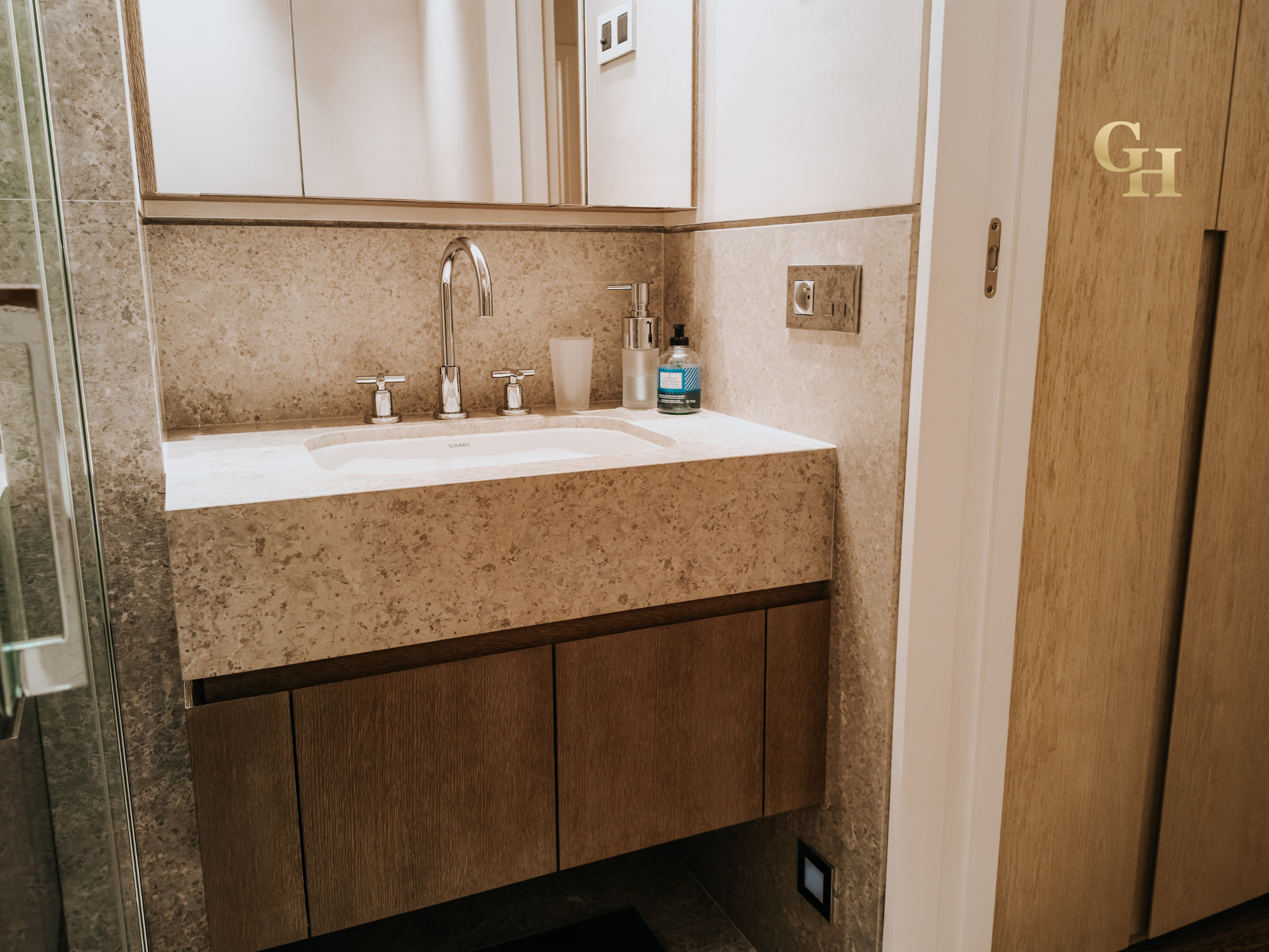 Bathroom sink and countertop with soap dispenser, a white cup, and a bottle of hand sanitizer, with mirrored medicine cabinet and wall outlet.
