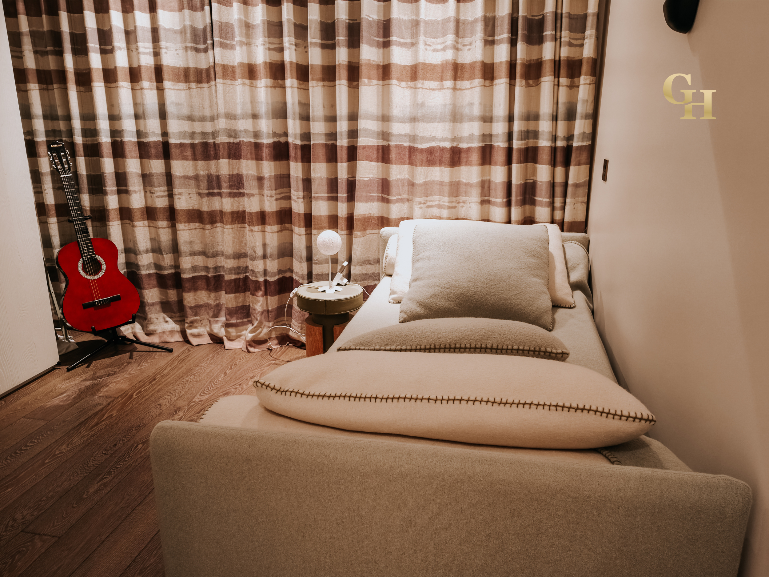 A cozy room with a bed, beige pillows, a lamp on a small side table, beige curtains with brown stripes, a red acoustic guitar leaning against the wall, and a wall decoration with the initials G and H.