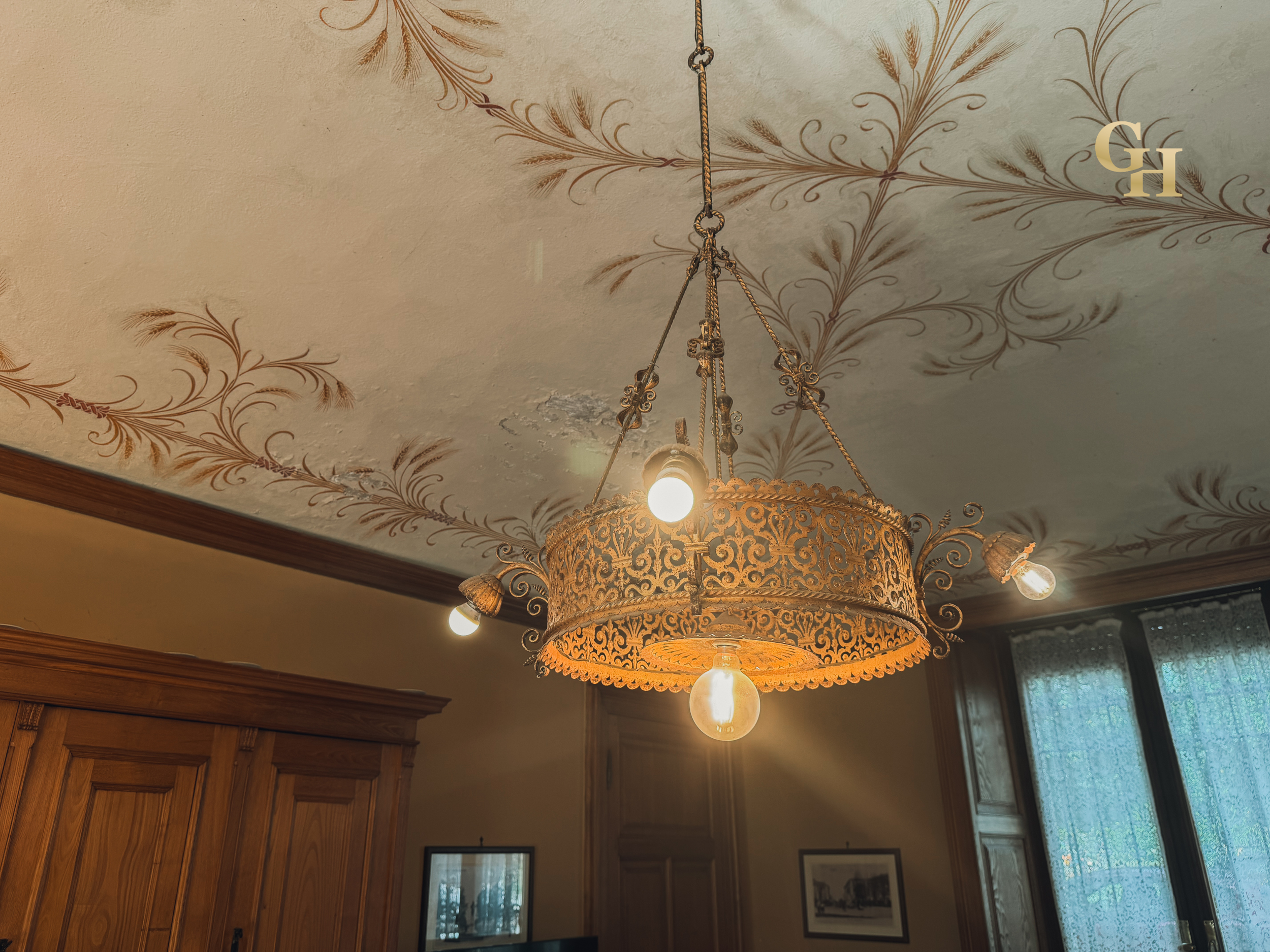 A close-up of a vintage hanging chandelier with ornate metalwork, four outward-facing bulbs, and intricate design, mounted on a ceiling decorated with painted gold-colored floral patterns.
