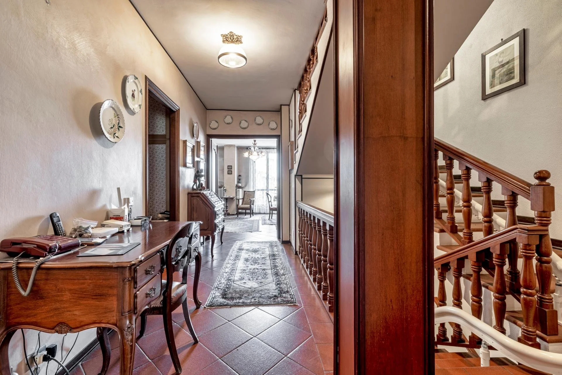 A hallway in a house with a wooden desk and chair on the left, decorated plates on the wall, and a carpet runner on the tiled floor. A staircase with wooden balusters on the right leads upstairs, and at the end of the hallway is a sitting area with c