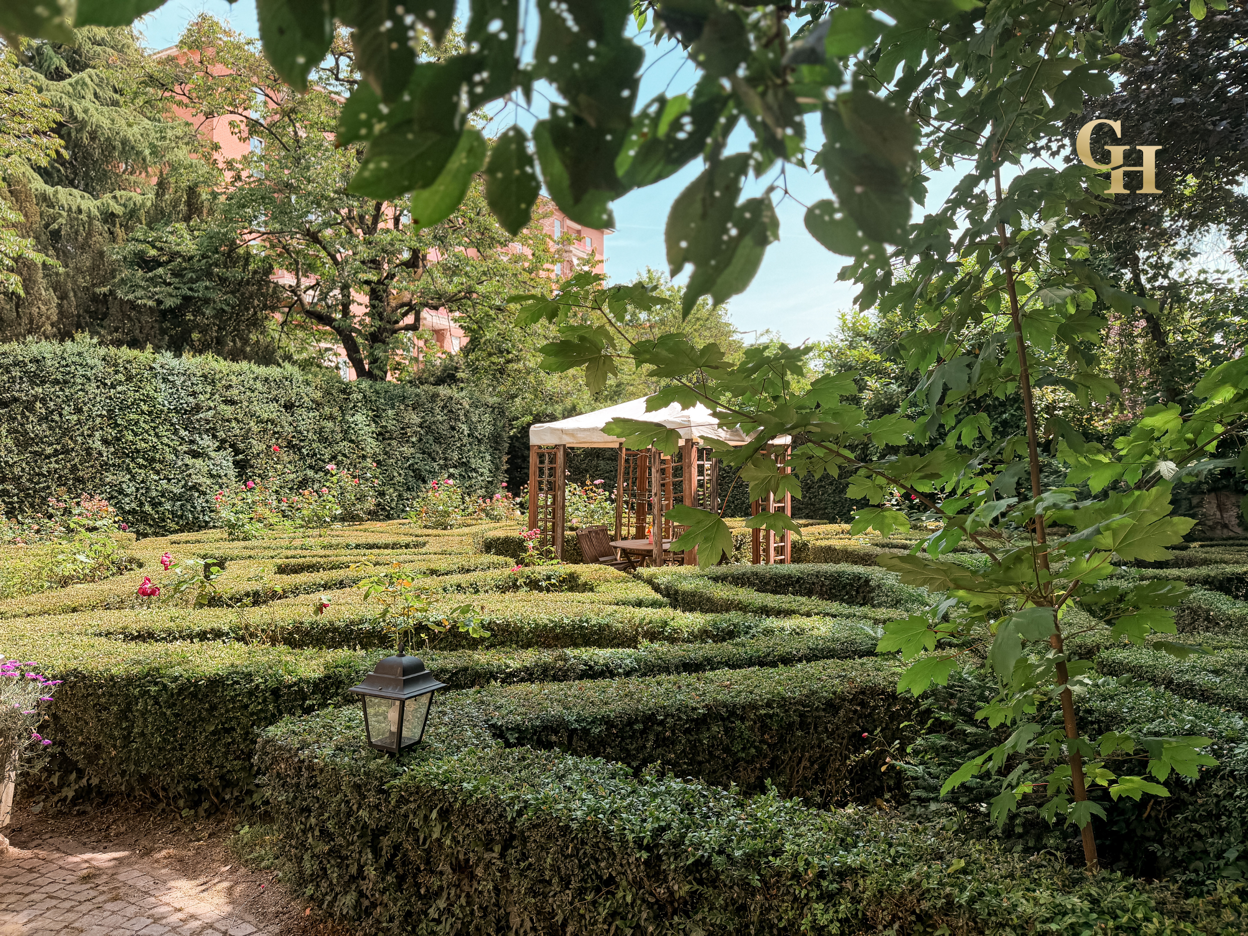 A lush garden with a variety of plants, flowers, and trees. A white canopy tent, benches underneath, and a brick building are visible in the background. The image is framed by green leaves in the foreground, with a small pathway and a lantern-style l
