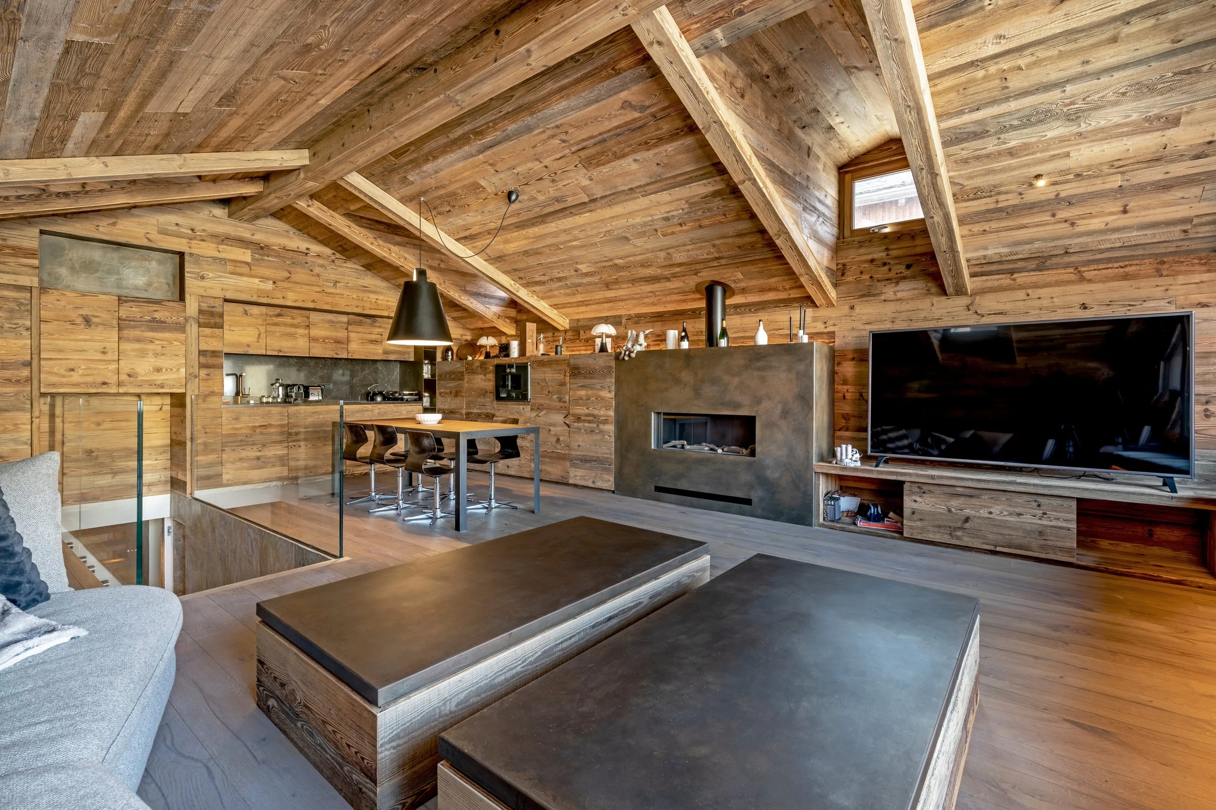 Interior view of a modern wood-paneled living room and kitchen with a high, angled ceiling, black hanging pendant light, flat-screen TV, and a kitchen island with bar stools.