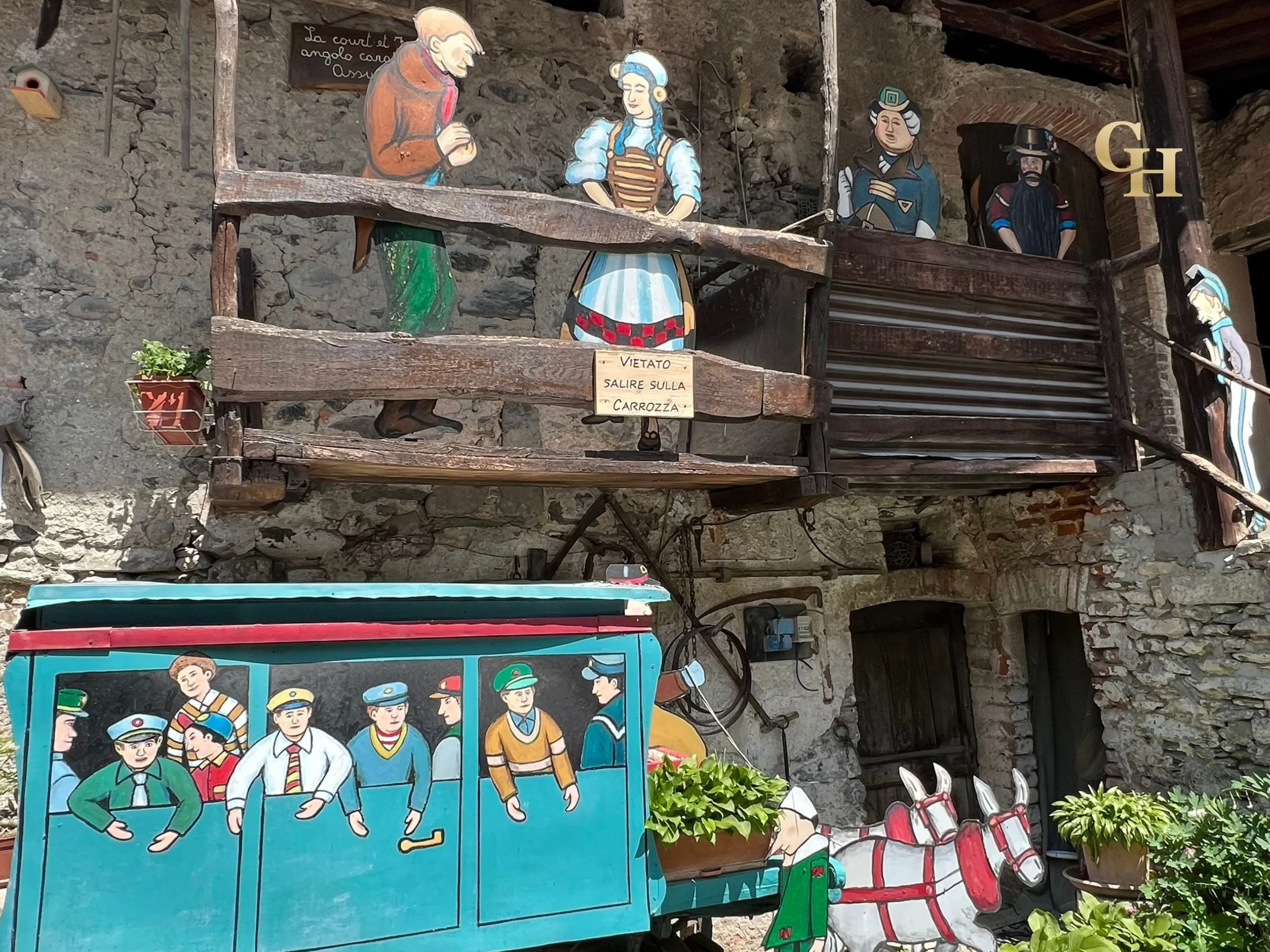 Colorful wooden decorations and figures depicting a historical or traditional scene, with a sign in Italian that says 'Vietato salire sulla carrozza', placed against a stone wall.