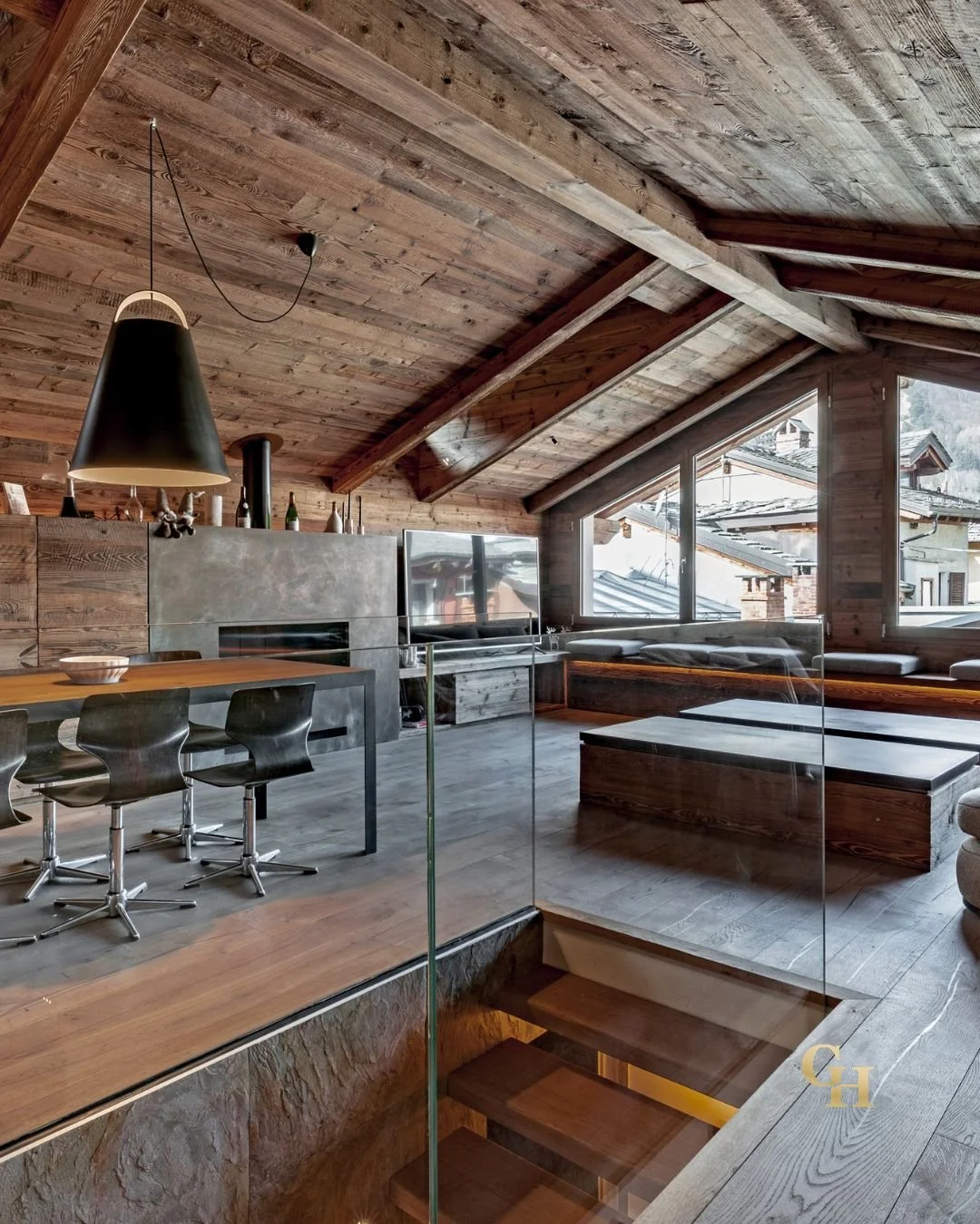 Modern attic living room with wooden walls, sloped ceiling, large windows showing snowy outdoor scenery, a flat-screen television, and a seating area with cushions.