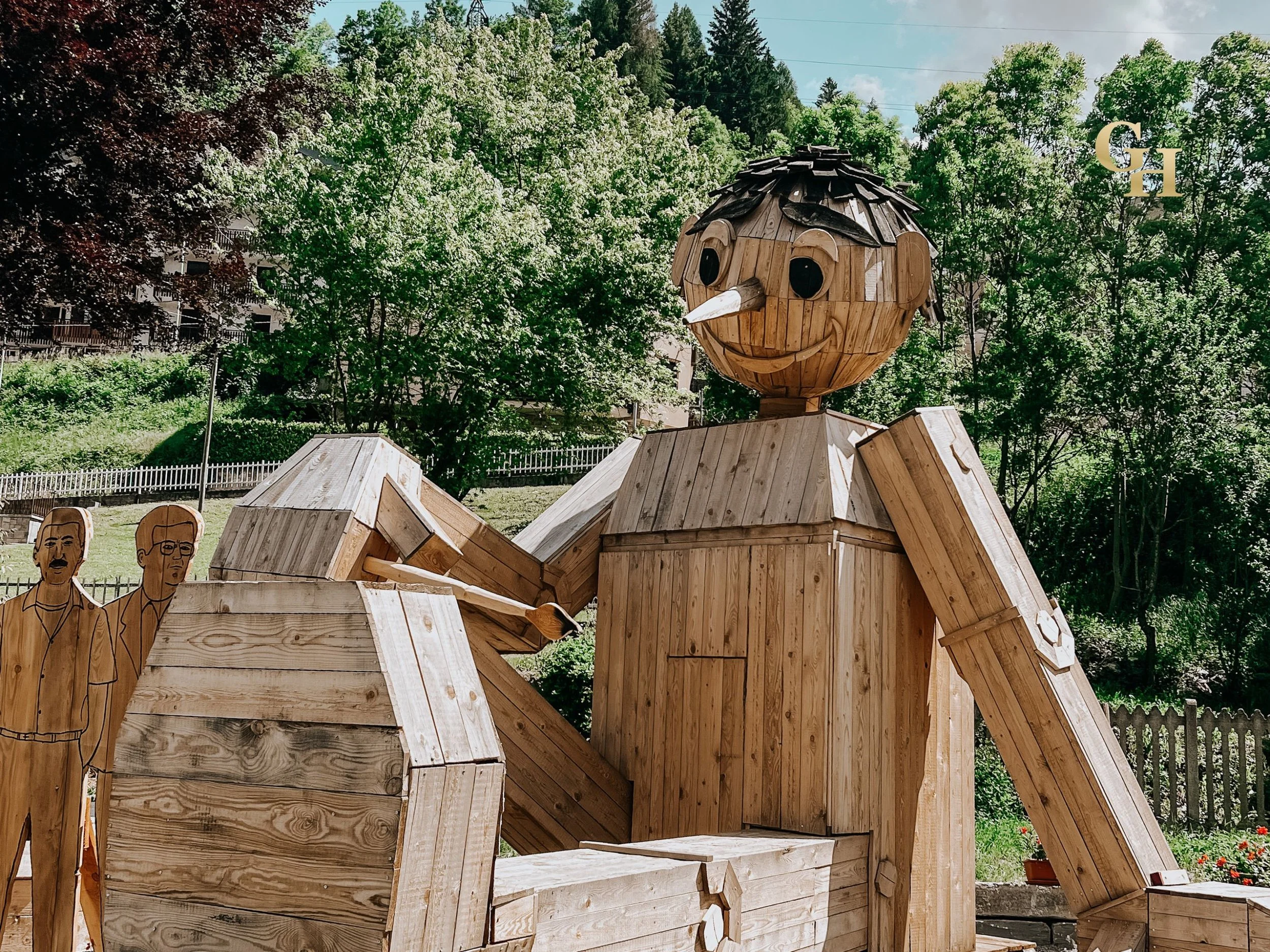 A large wooden sculpture of Pinocchio with a smiling face, black eyes, a long nose, and dark hair. The sculpture is outdoors near trees and has a decorative gold and black shield with the letter 'G' and 'H' in the background.