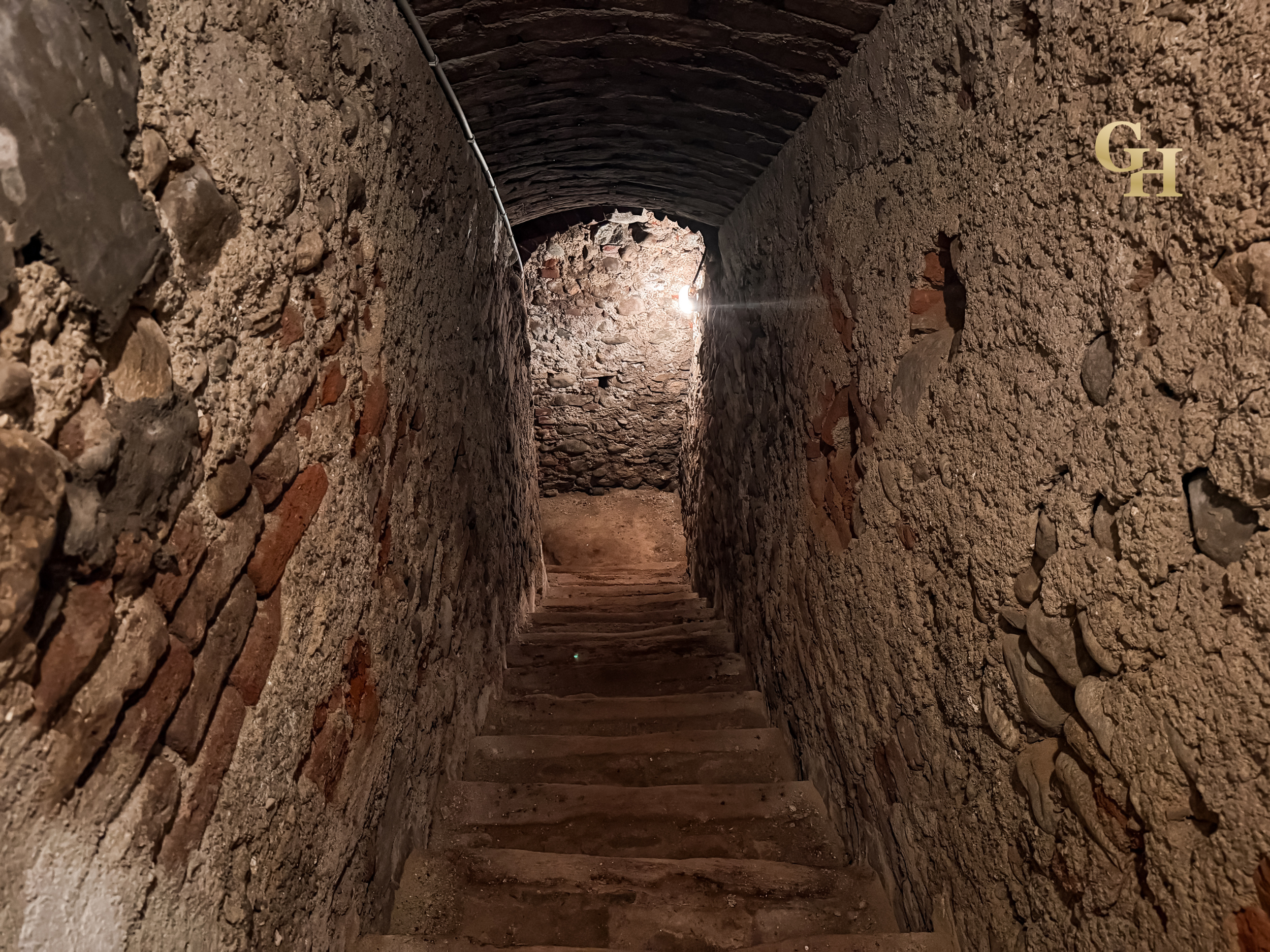 A narrow underground tunnel made of stone with a curved ceiling and wooden steps ascending toward light at the end.