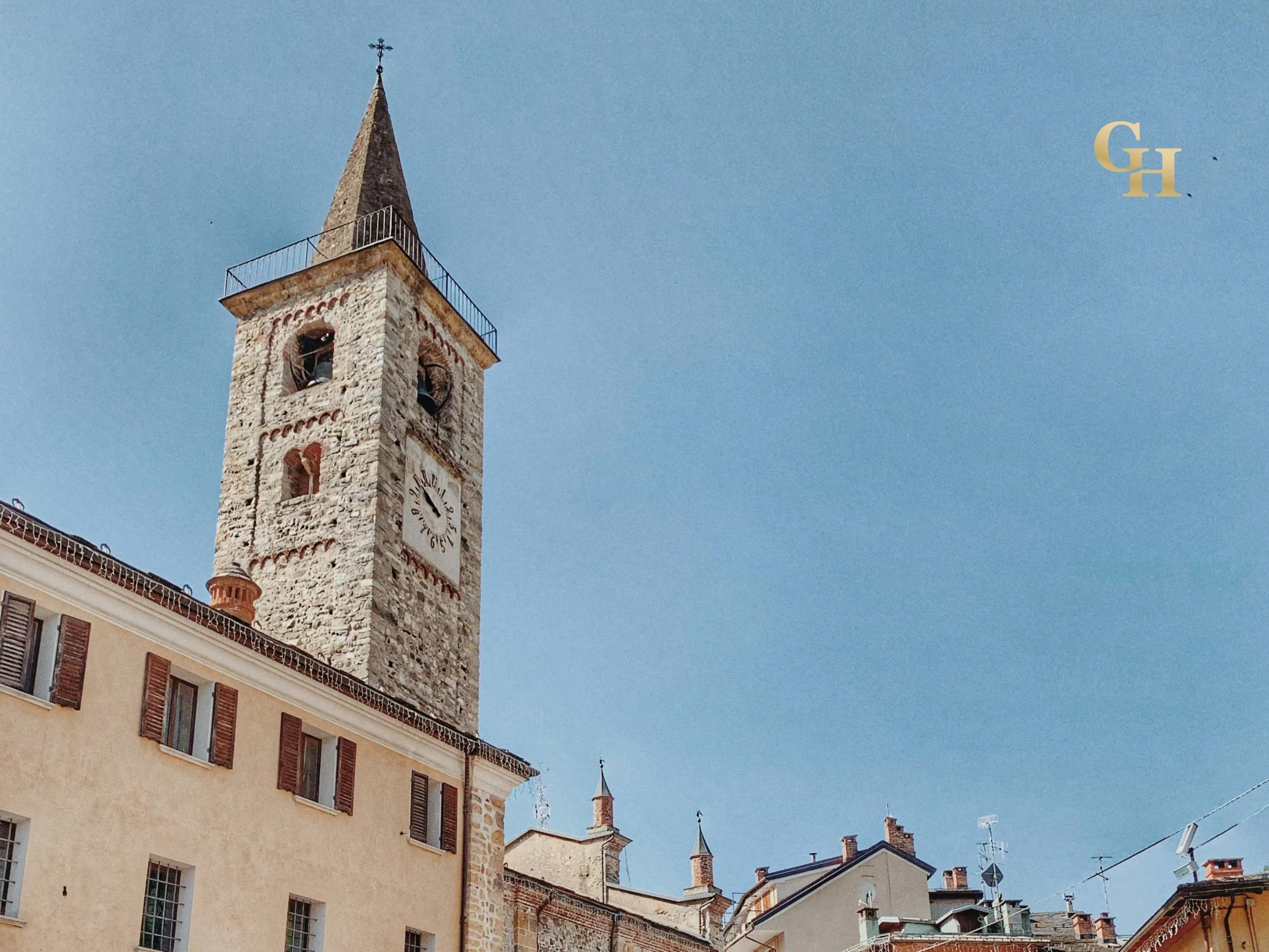 A stone clock tower with a spire and weather vane, set against a clear blue sky, with surrounding buildings with multiple stories and chimney stacks. Historic centre of Limone Piemonte, Piedmont, Italy.