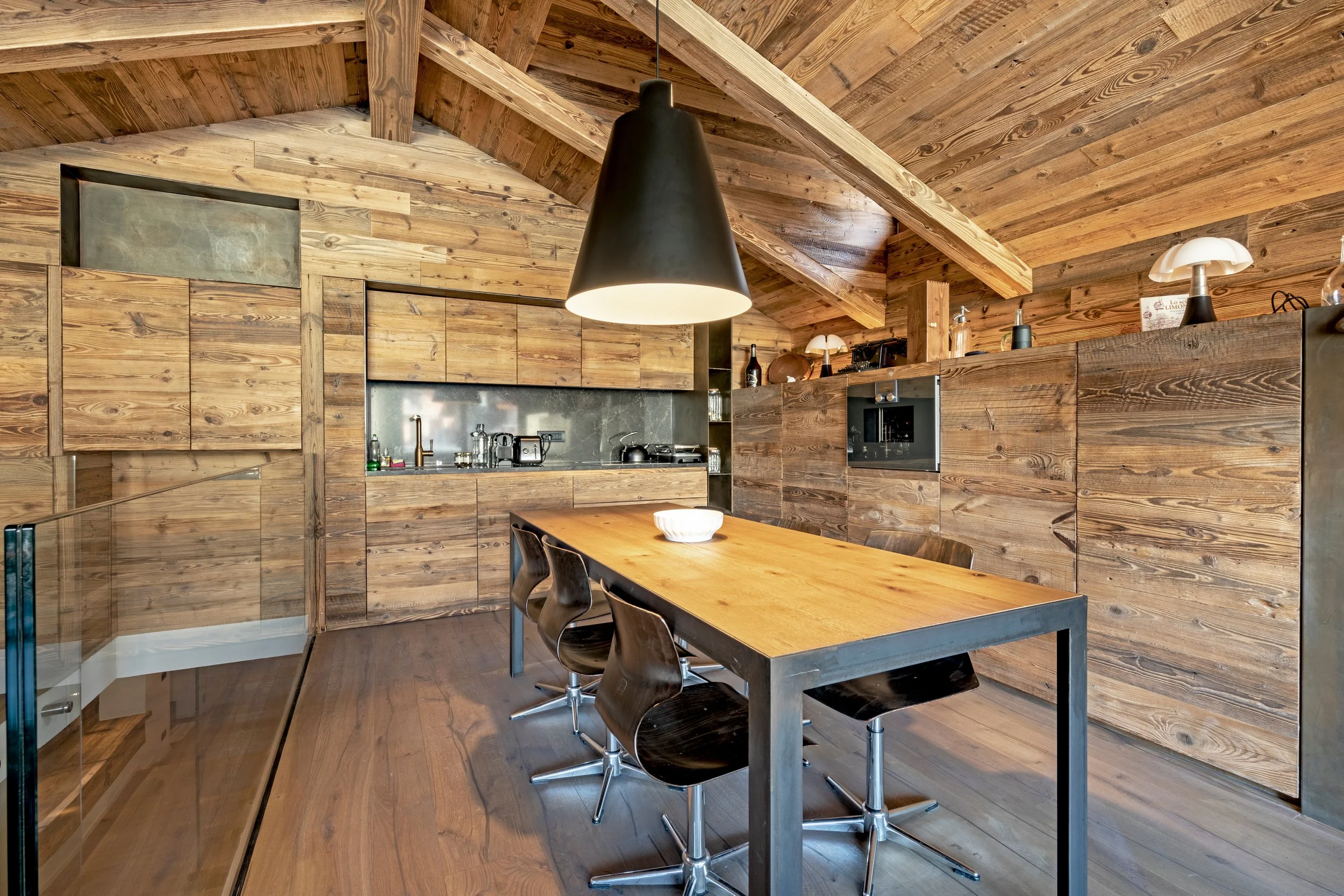 A modern kitchen with wooden paneling and ceiling, featuring a wooden dining table with black chairs, black pendant light, and various kitchen appliances and utensils.