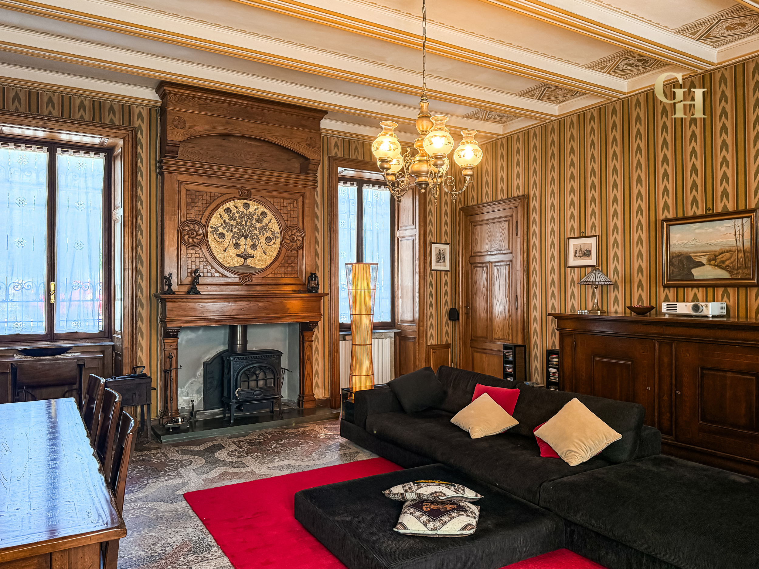 Living room with wood paneling, cabinets, a fireplace, artwork on the walls, a chandelier, large windows with lace curtains, a black sofa with pillows, a red carpet, and a wooden dining table with chairs.