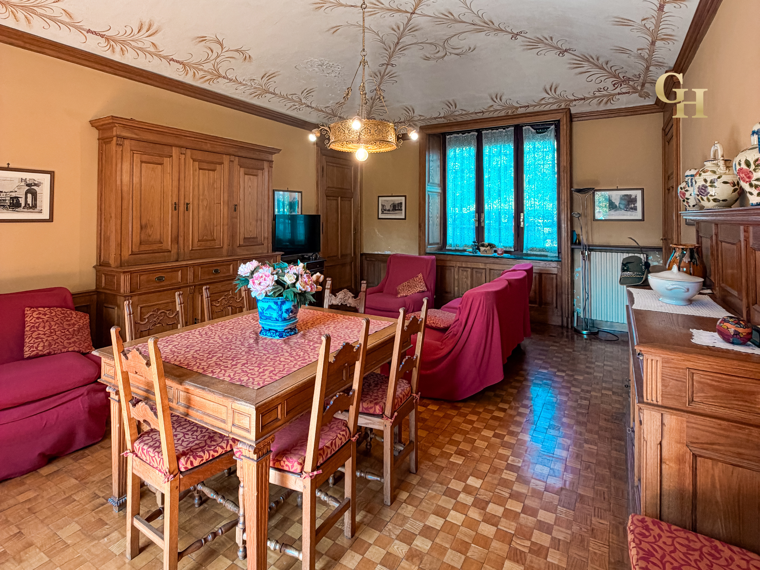 A cozy living room with wooden furniture, red upholstered chairs, a dining table covered with a patterned tablecloth, and a decorated ceiling with floral designs. There is a plant on the window sill, a television, and decorative vases on a cabinet.
