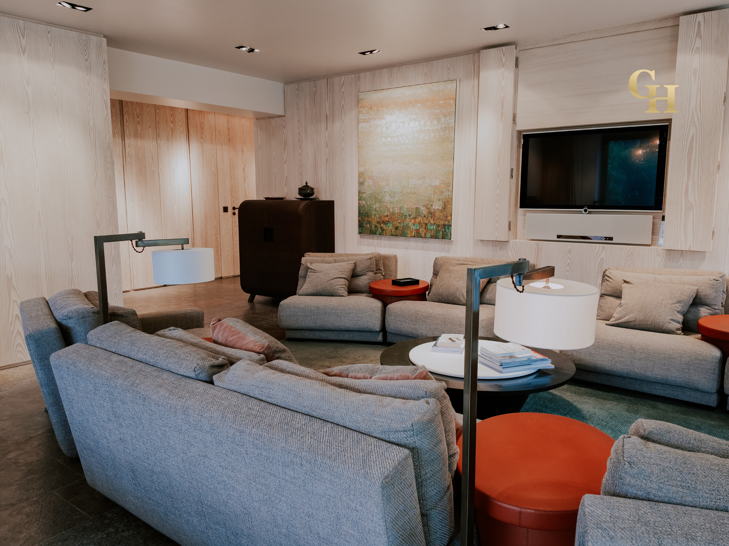 A modern living room with gray sofas, orange side tables, a large abstract painting on the wall, a flat-screen TV, and a wooden TV stand, with wood-paneled walls and ceiling lights.