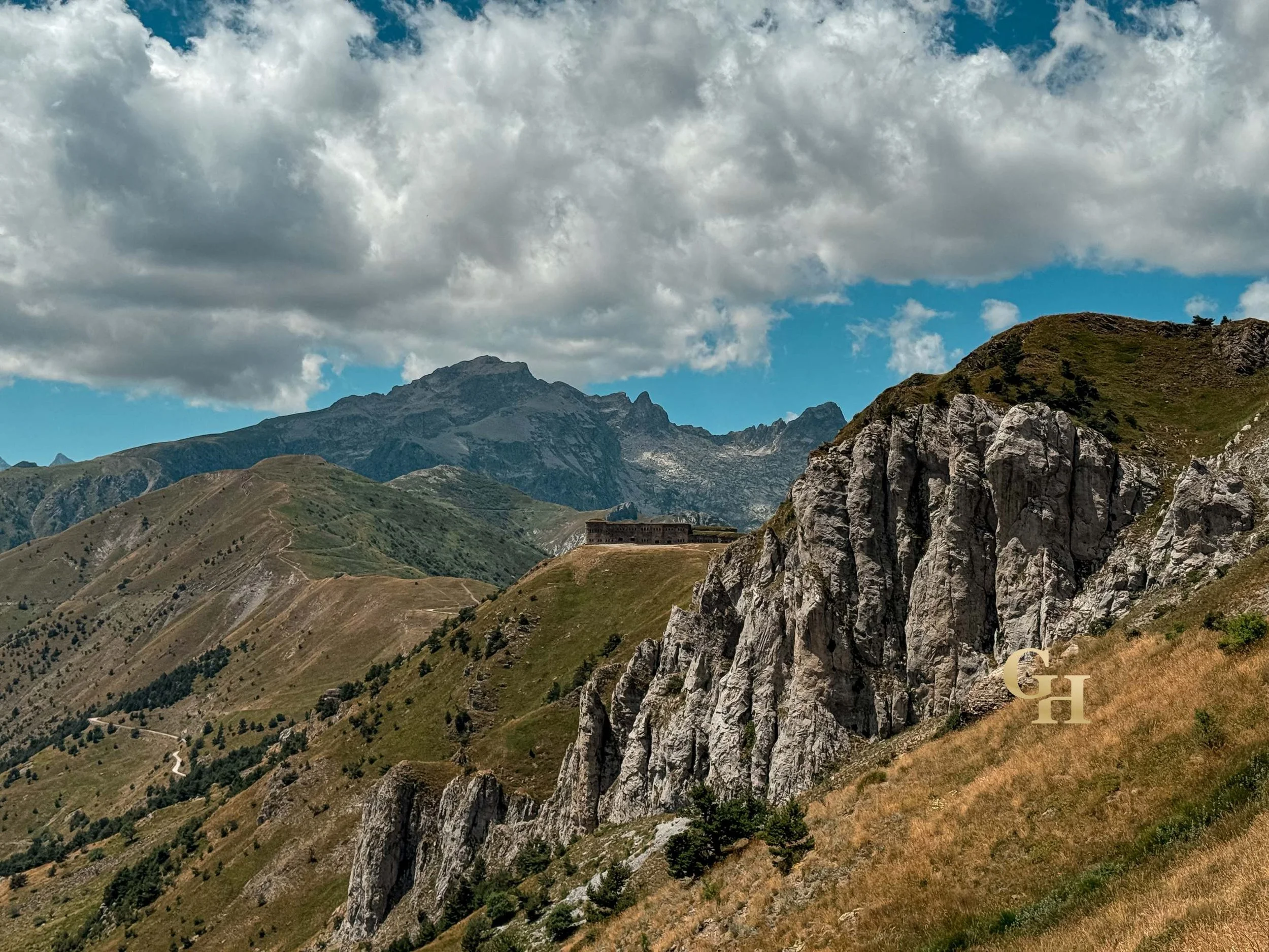 A scenic mountain landscape with green hills, rocky cliffs, a building, and a cloudy sky. Landscape of the Maritime Alps, Piedmont, Italy