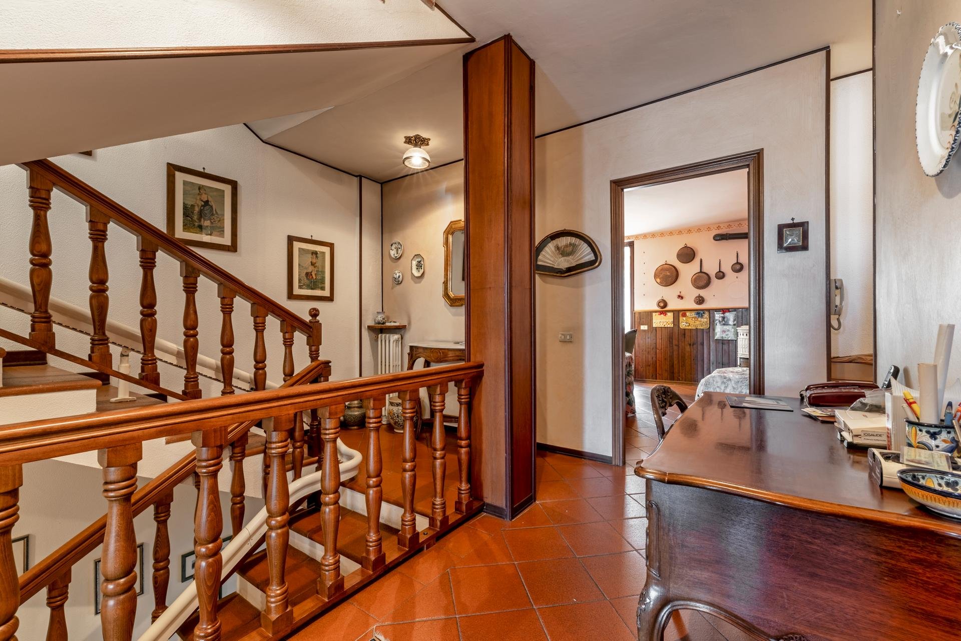 Interior view of a house with a curved staircase with wooden railing, adjacent to a dining area with a wooden table and chairs, and a doorway leading to a living room decorated with hanging pots and artwork.