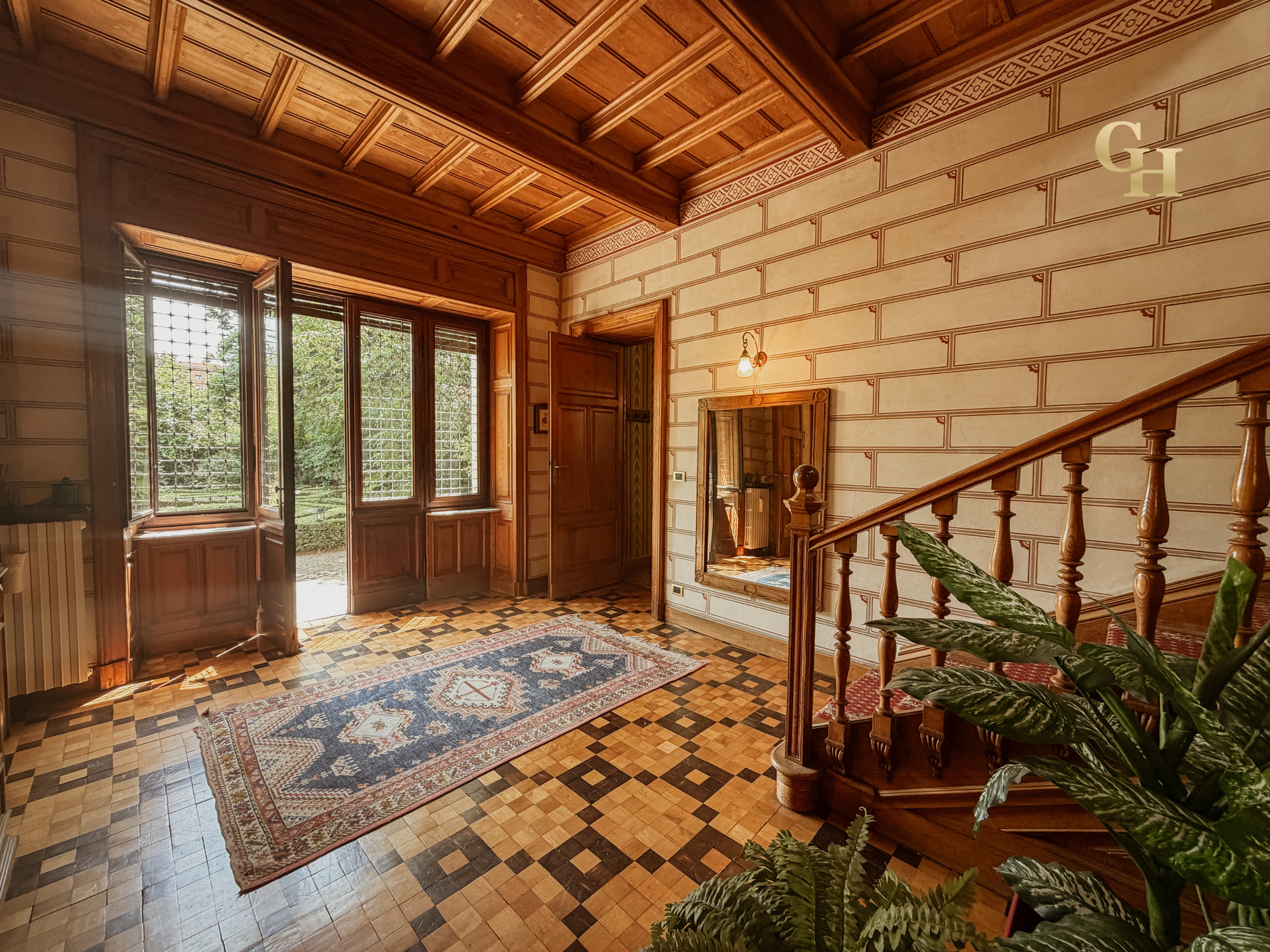 Interior view of a wooden staircase and hall with a decorative rug, large windows with blinds, and a mirror on the wall.