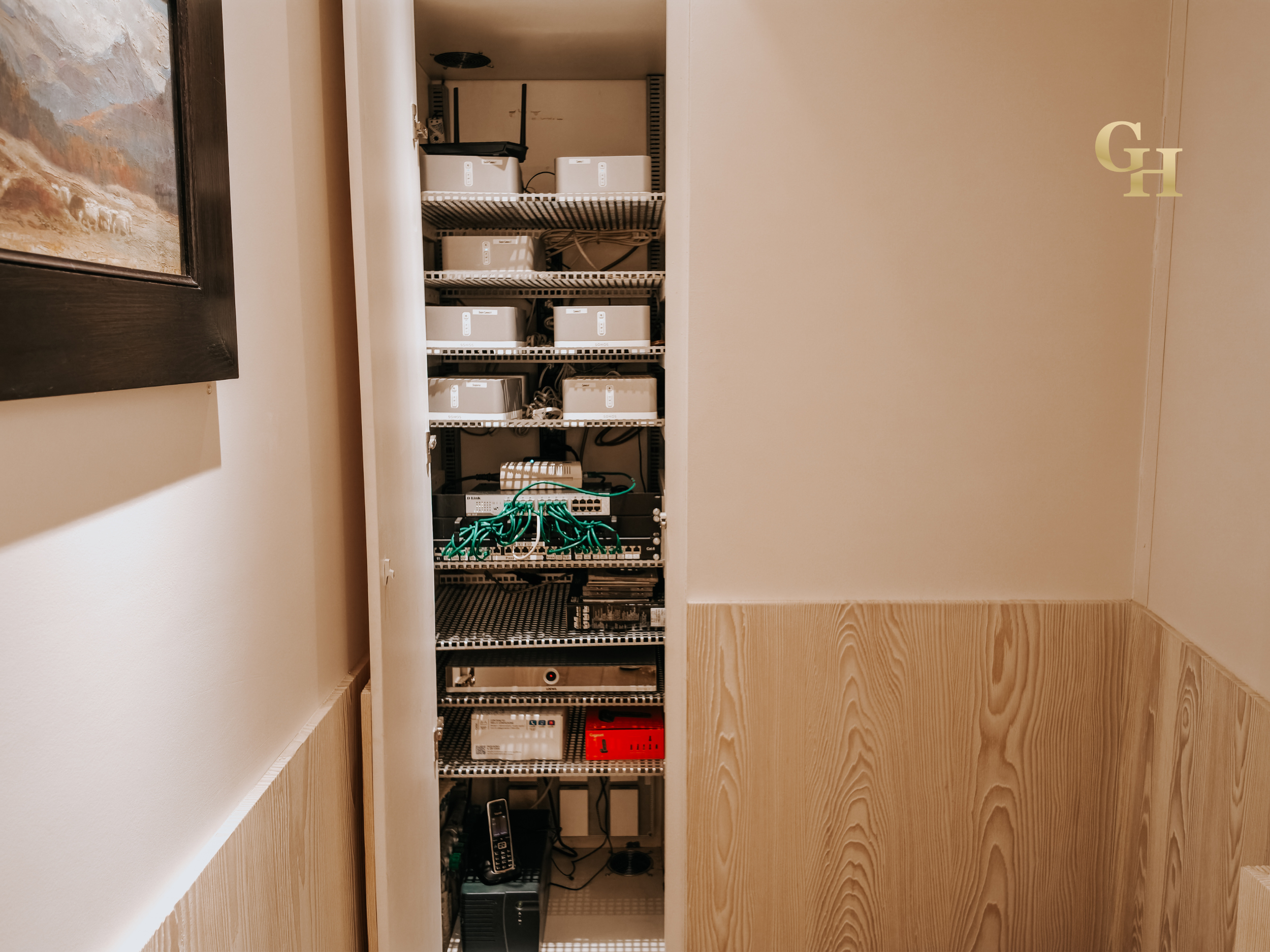 A tall, open server or networking equipment rack with multiple components, including switches, routers, and power supplies, placed against a wall with a framed painting on the left and a wooden panel on the right.