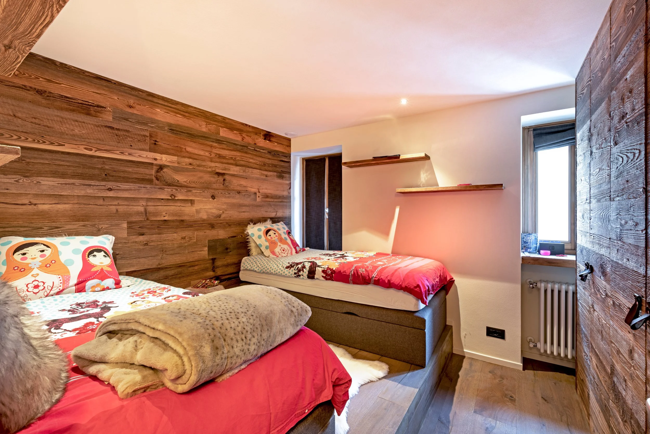 A cozy bedroom with a wooden accent wall, two beds with colorful bedding and cartoon-themed pillows, a window with blinds, and floating shelves on a beige wall.