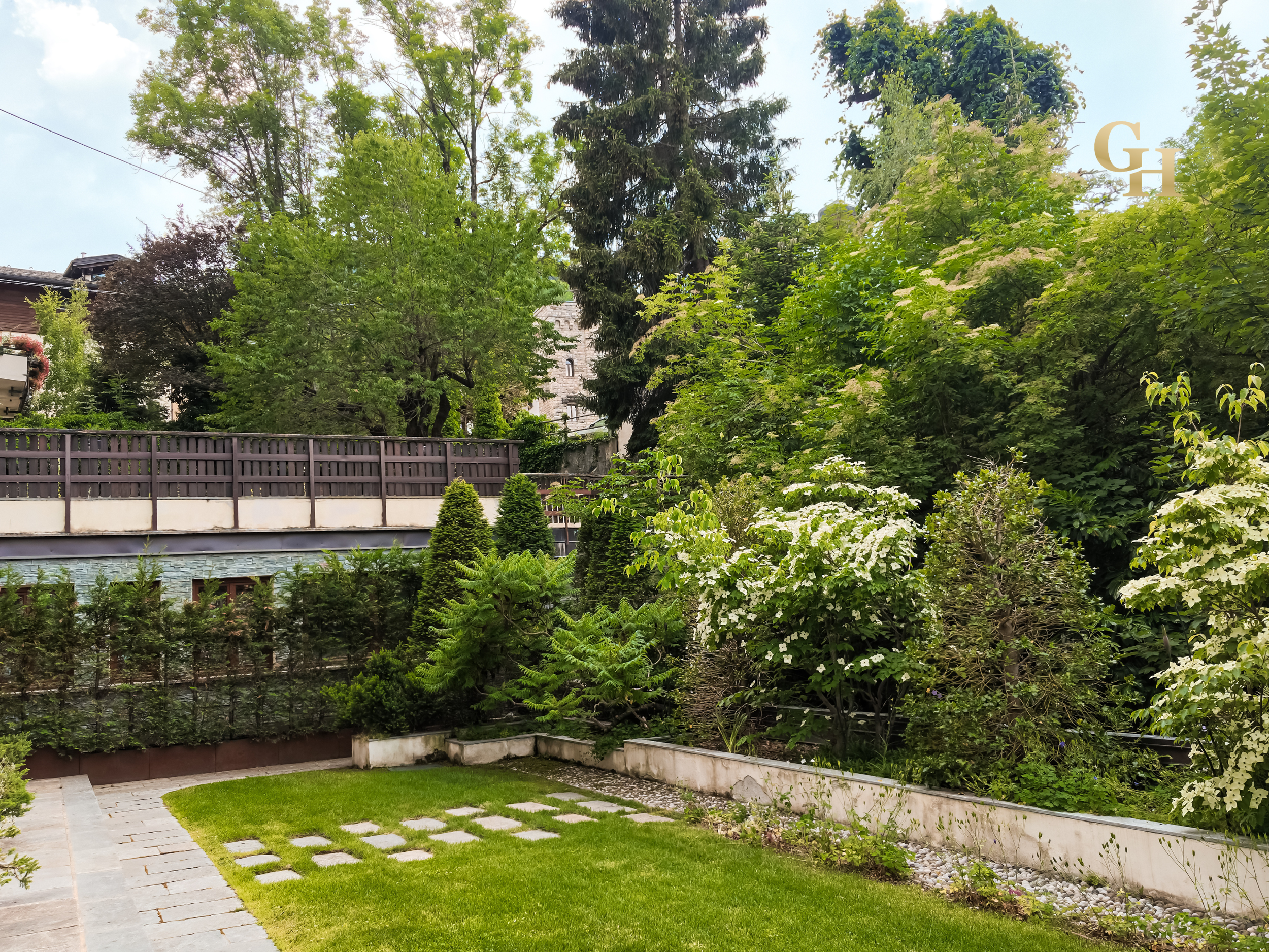 A garden with a well-maintained grass lawn and stepping stones, surrounded by a variety of lush green plants, small trees, and shrubs, with a taller tree and buildings visible in the background.